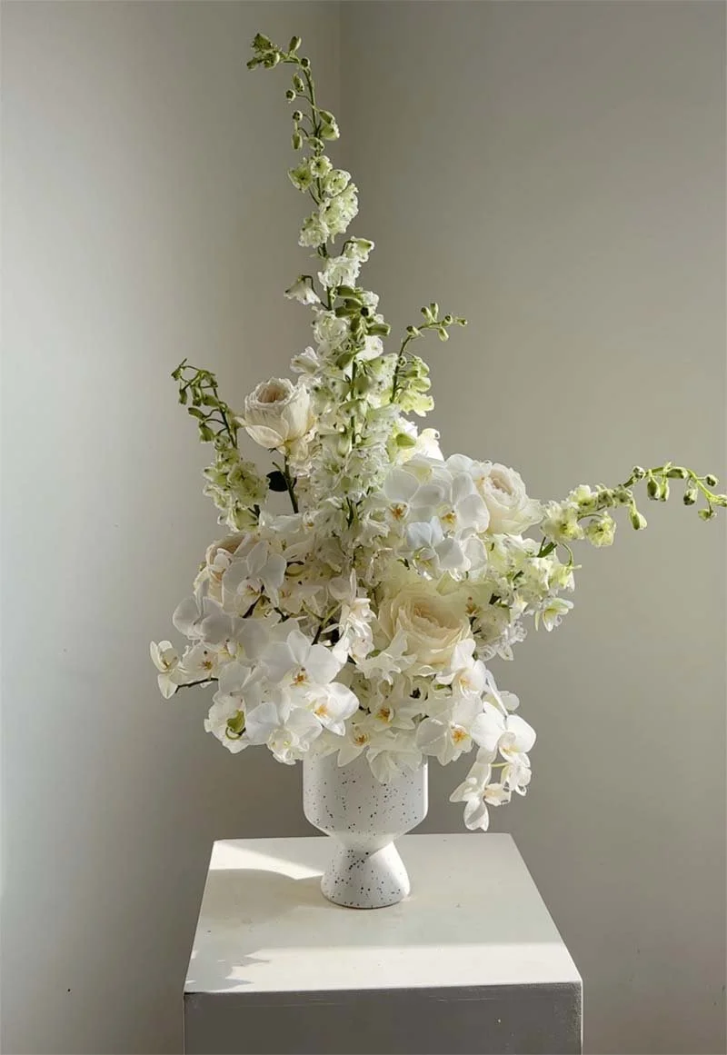 Elegant white flower arrangement with orchids and roses in a white speckled vase on a white pedestal.