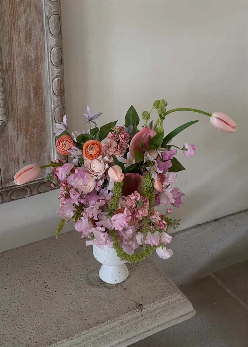 Pink, peach, and purple flowers in a white vase on a stone surface, with a wooden-framed mirror in the background.