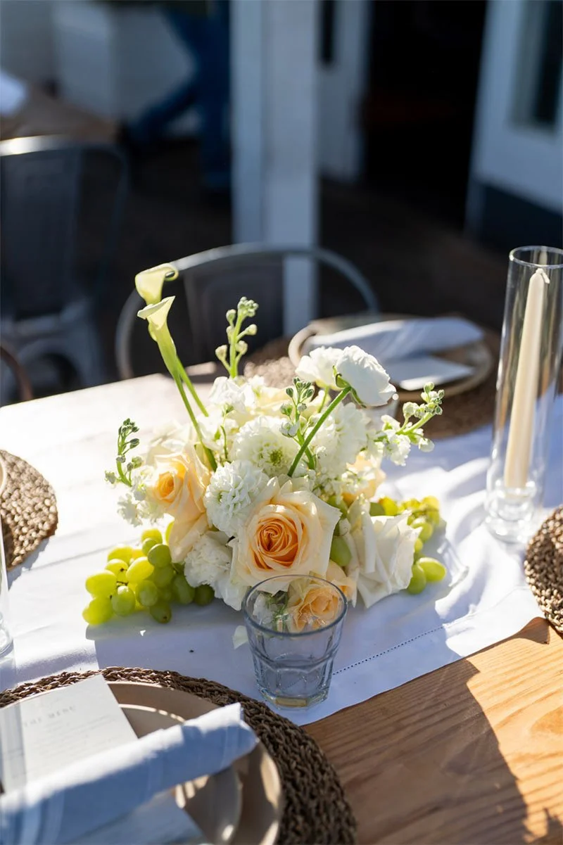 A floral centerpiece with white and peach flowers and green grapes on a table set for a meal, including a glass with a peach rose, a tall candle, and place settings with napkins and plates.