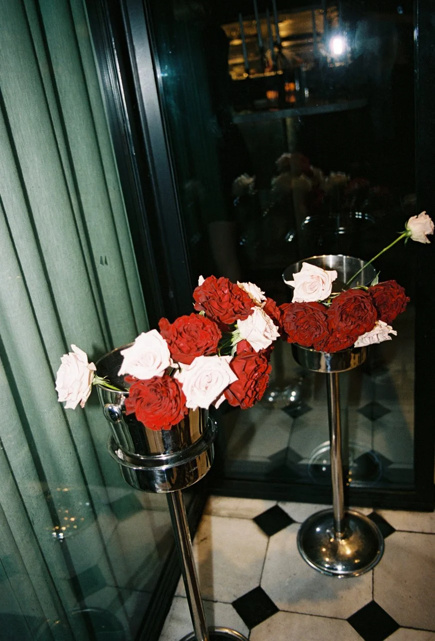 Two metallic flower stands with artificial roses in red and white, placed indoors near a glass wall, with a tiled floor and reflections on glass.