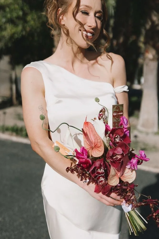 A woman in a white dress holding a bouquet of pink, purple, and green flowers outdoors, smiling with her eyes closed.