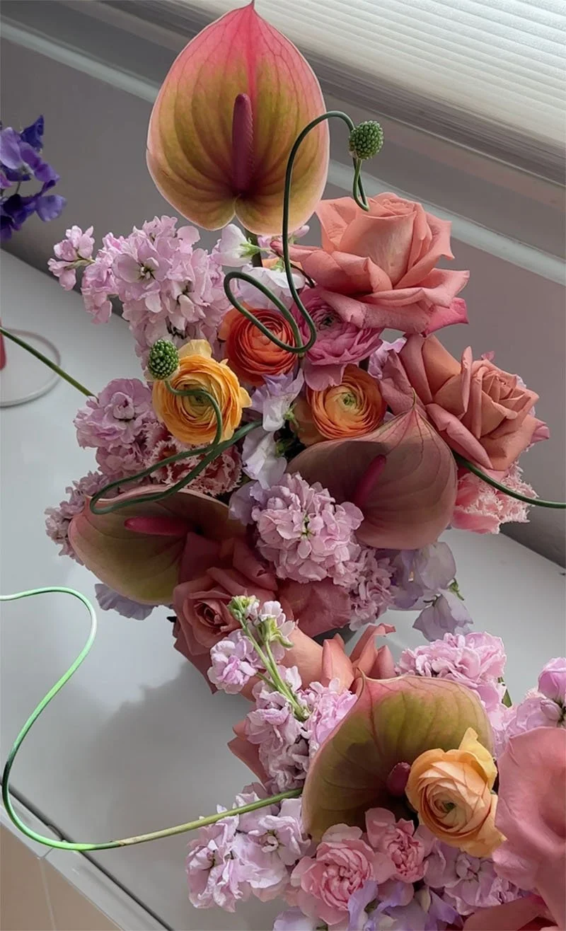 A bouquet of pink and peach roses, calla lilies, and ranunculus flowers in a vase with wire spirals, arranged on a white surface near a window.