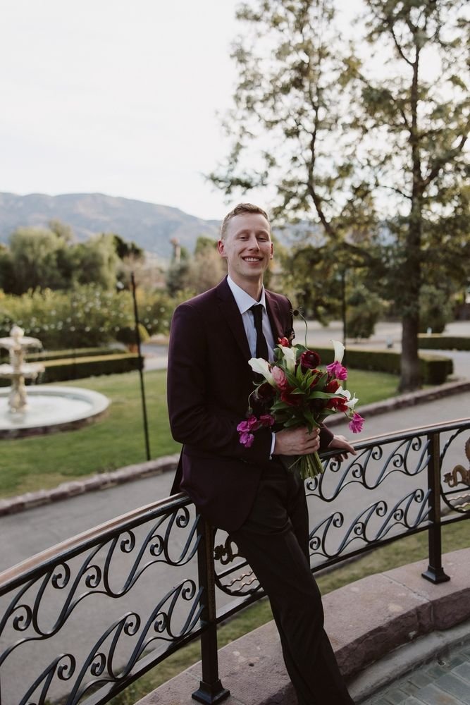 A smiling young man in a black suit holding a bouquet of flowers on a balcony outdoors.