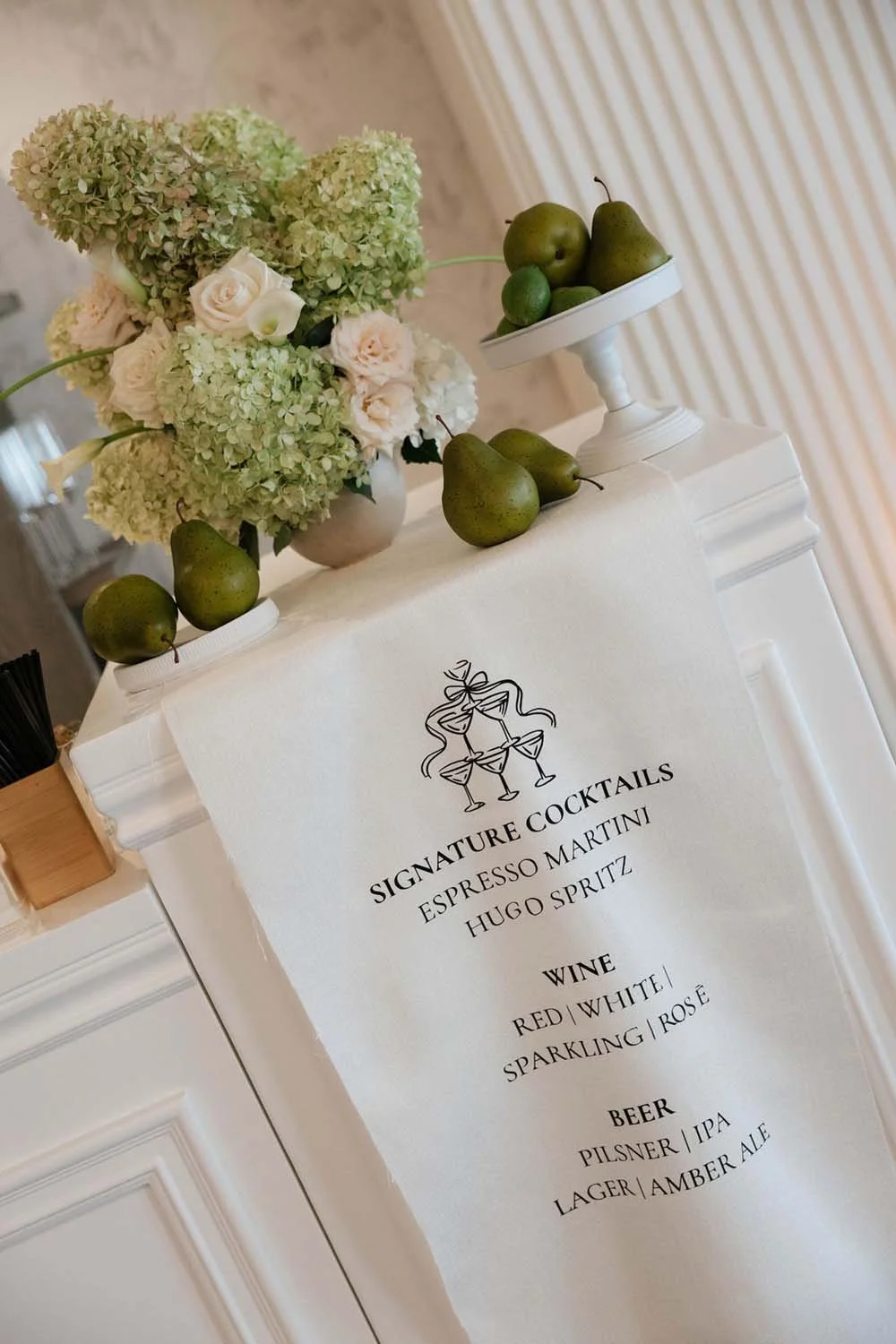 Elegant table display with a floral arrangement of hydrangeas and roses, a tray of green pears, and a sign listing signature cocktails, wine, and beer options.