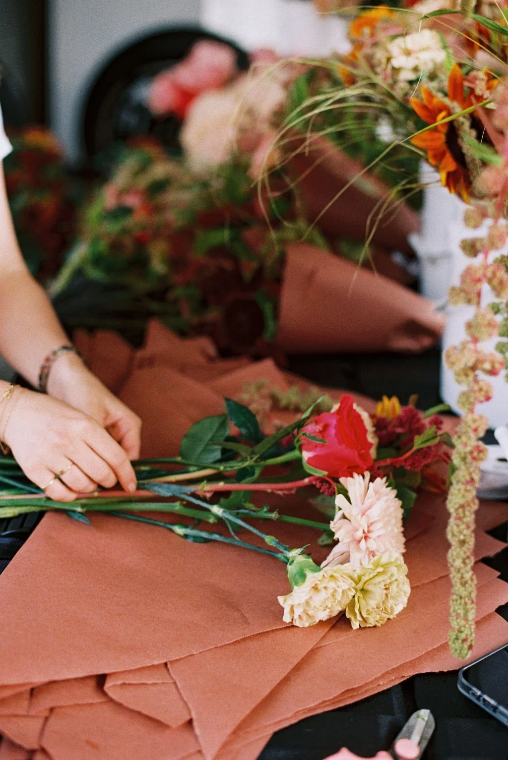 Person's hands arranging pink, red, and yellow flowers on brown craft paper with a blurred background of more flowers.
