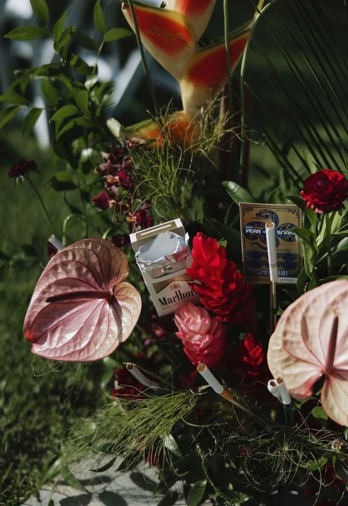 A floral arrangement with pink and red flowers, various greenery, and a large tropical leaf with a red hue. There are also cigarette packs, a matchbox, and a cigarette in a bright outdoor setting.