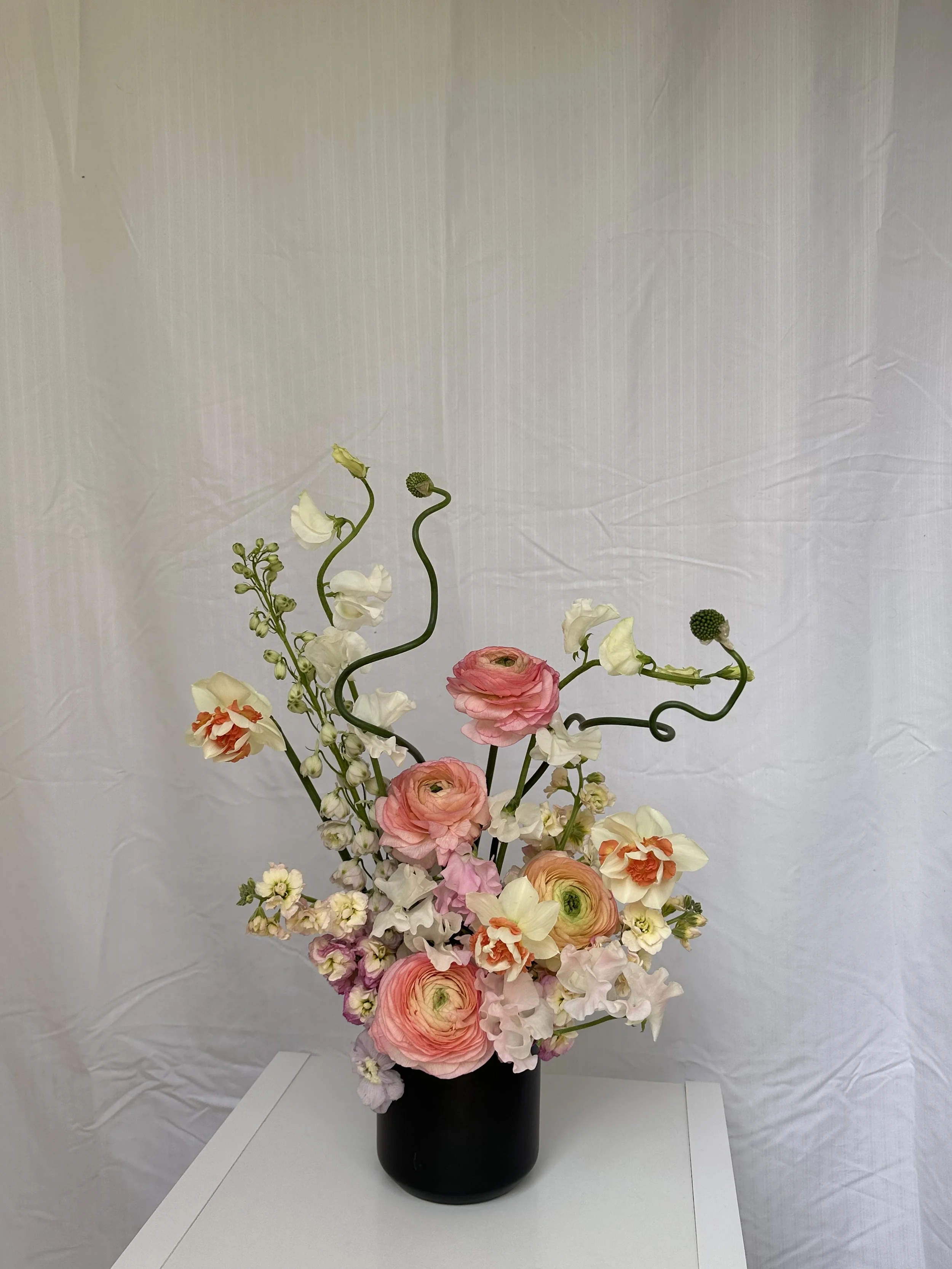 A floral arrangement in a black vase with pink, white, and orange flowers and green curly stems on a white surface against a beige background.
