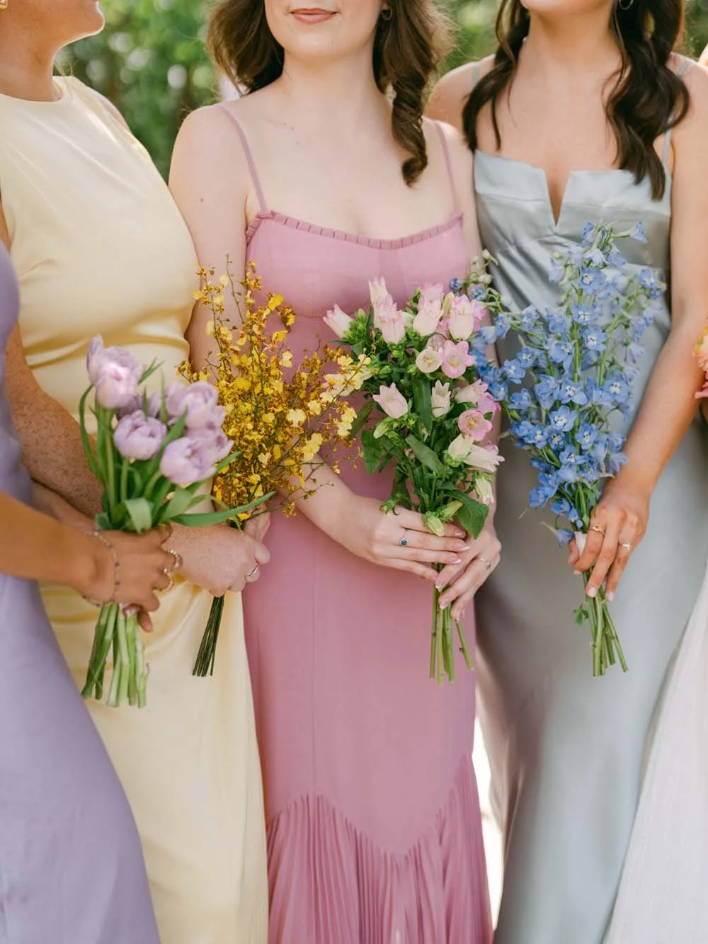 Close-up of four women holding bouquets of pink, purple, yellow, and blue flowers, dressed in pastel-colored dresses, outdoors in a garden setting.