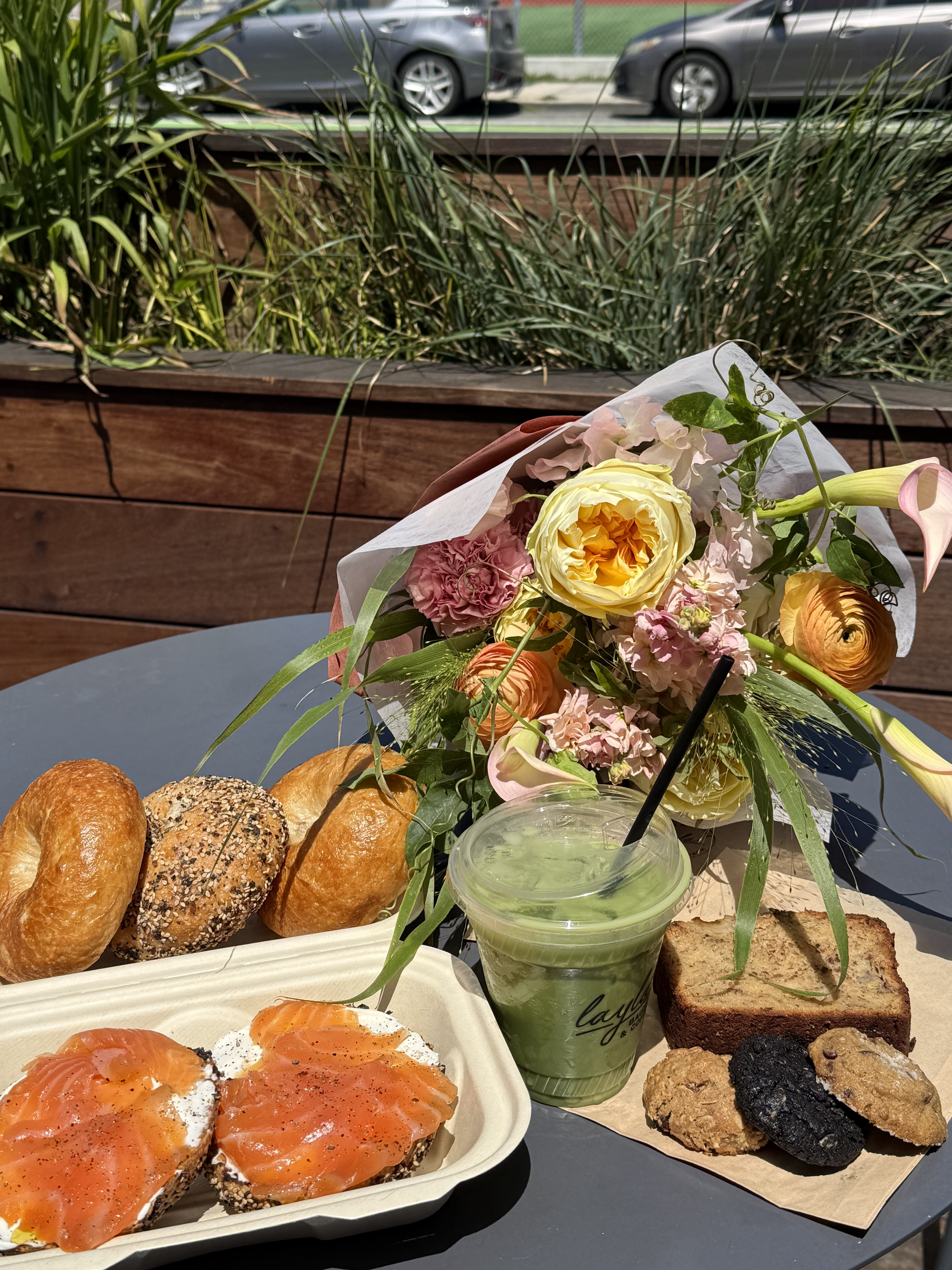 A tray of assorted bagels with smoked salmon on cream cheese, a green smoothie in a plastic cup, a slice of banana bread, and several cookies, all on a table outdoors with a bouquet of pink and yellow flowers in the background.