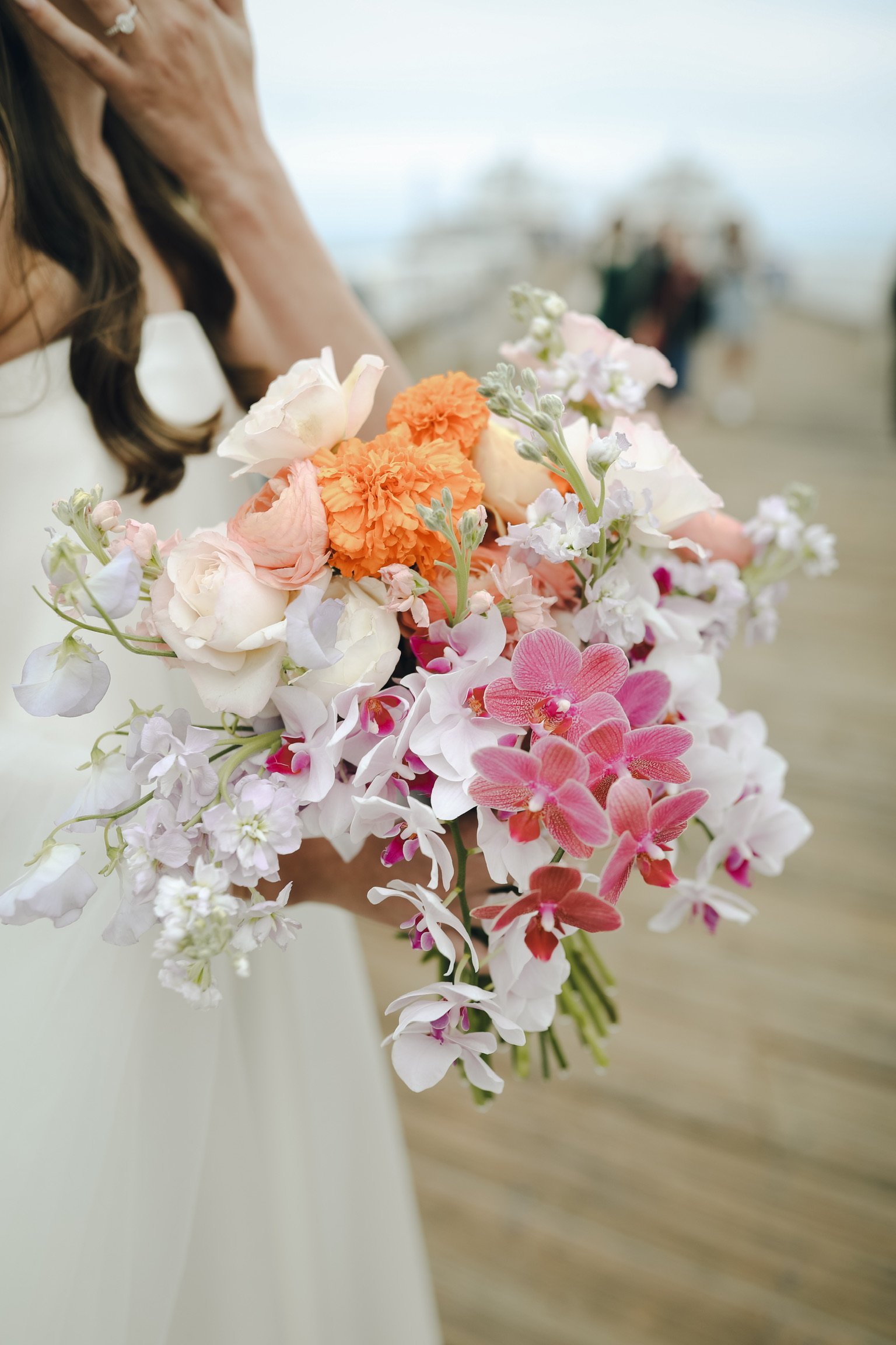 A woman in a white dress holding a large bouquet of various pastel-colored flowers, including pink orchids, orange carnations, and white roses, on a beach.
