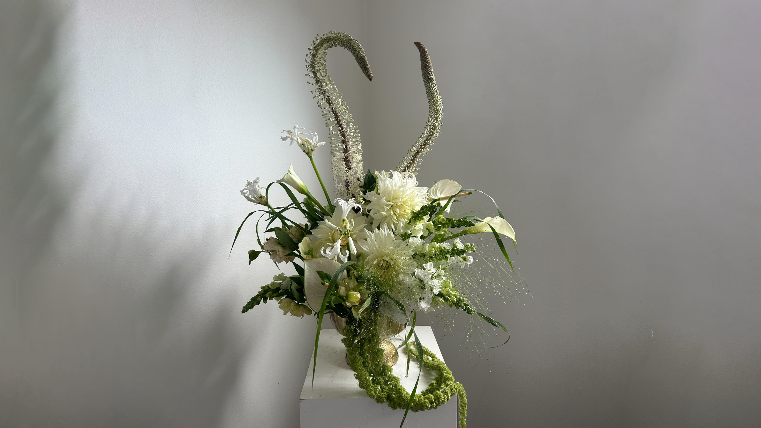 Elegant floral arrangement with white flowers and greenery on a white pedestal against a plain light gray wall.