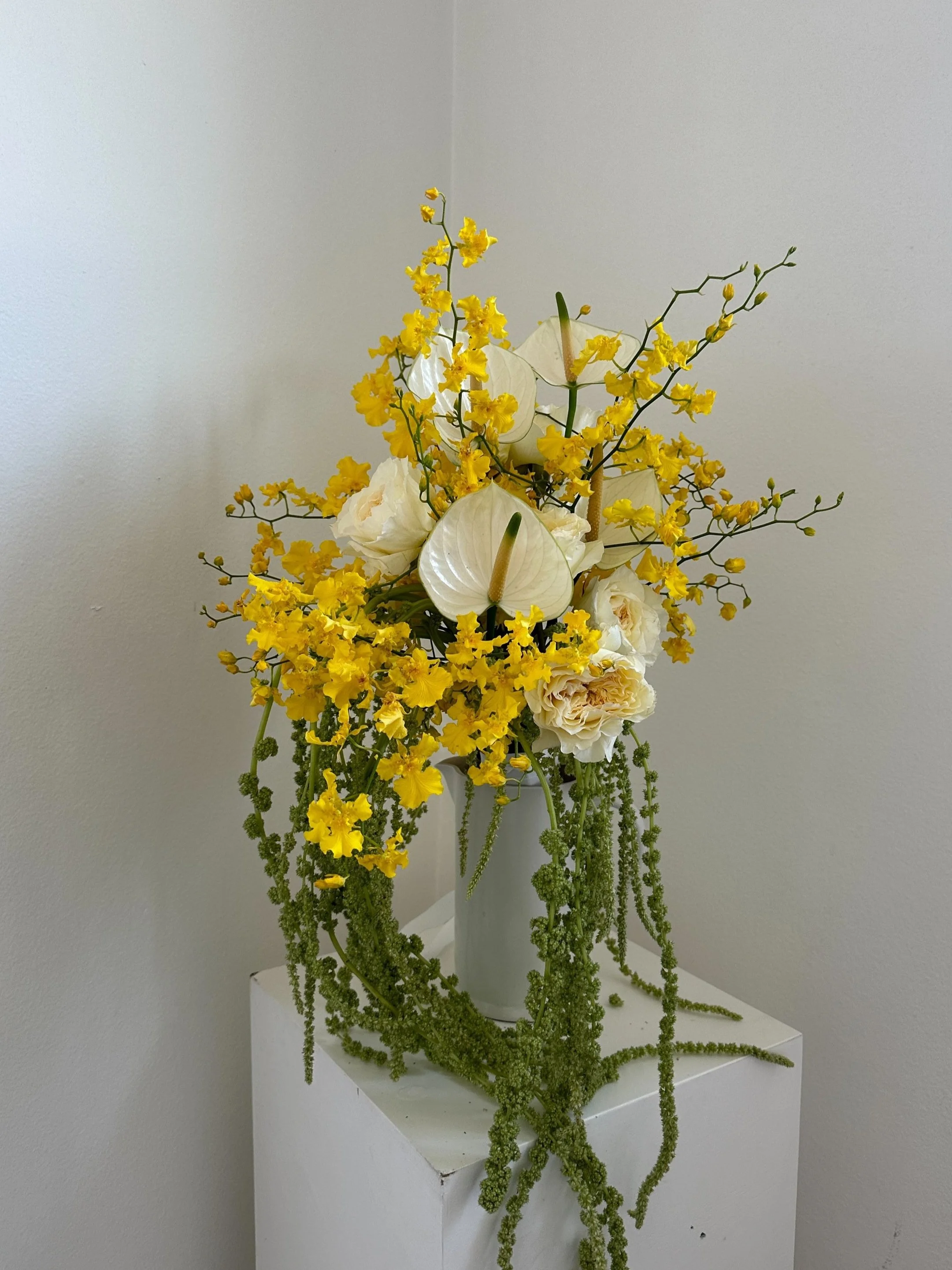 Yellow and white floral arrangement in a white vase on a white pedestal against a plain wall.