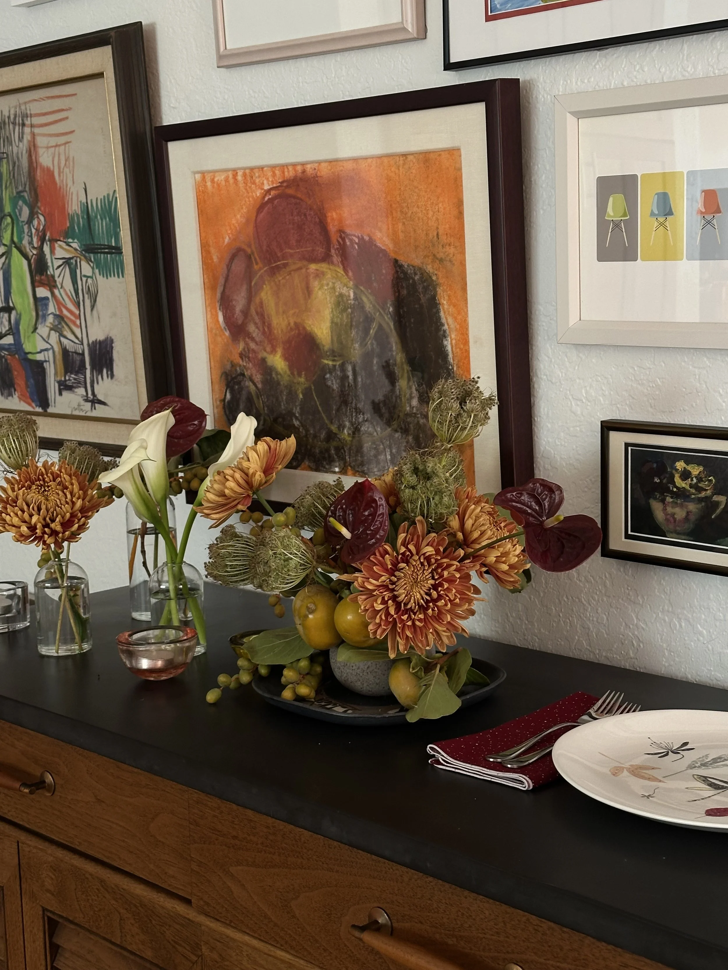 A floral centerpiece with orange, red, and green flowers and fruits on a black table, surrounded by art and framed pictures on the wall.
