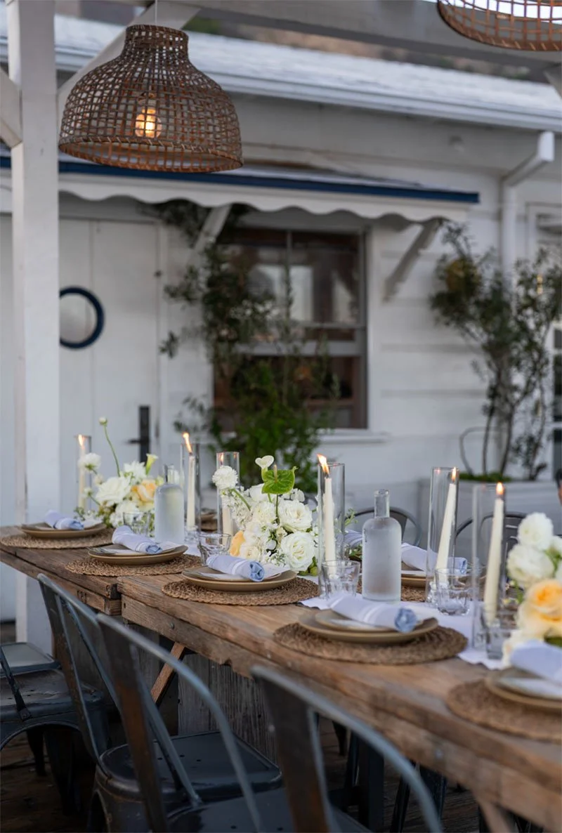 A rustic outdoor dining table decorated with white floral centerpieces, candles in tall glass holders, and place settings with beige plates and blue napkins, under woven pendant lights.