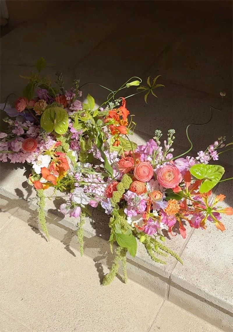 A colorful flower arrangement with pink, purple, orange, and green flowers arranged on a tiled surface with sunlight casting shadows.