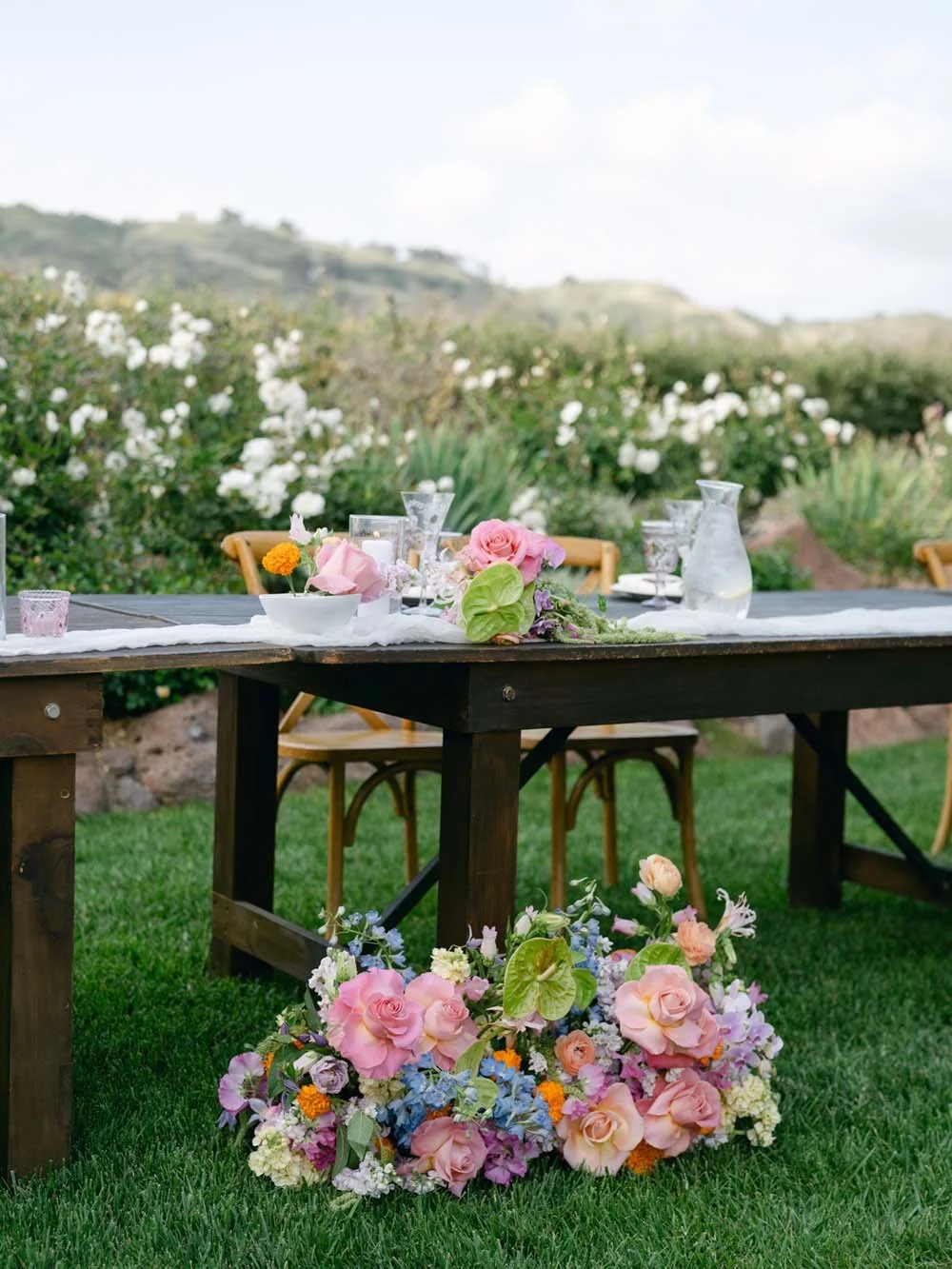 Outdoor table decorated with flowers, tableware, and glassware, set in a garden with greenery and hills in the background.