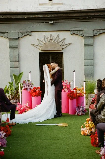 Bride and groom sharing a kiss during their wedding ceremony outside, with guests seated on either side and colorful floral arrangements.