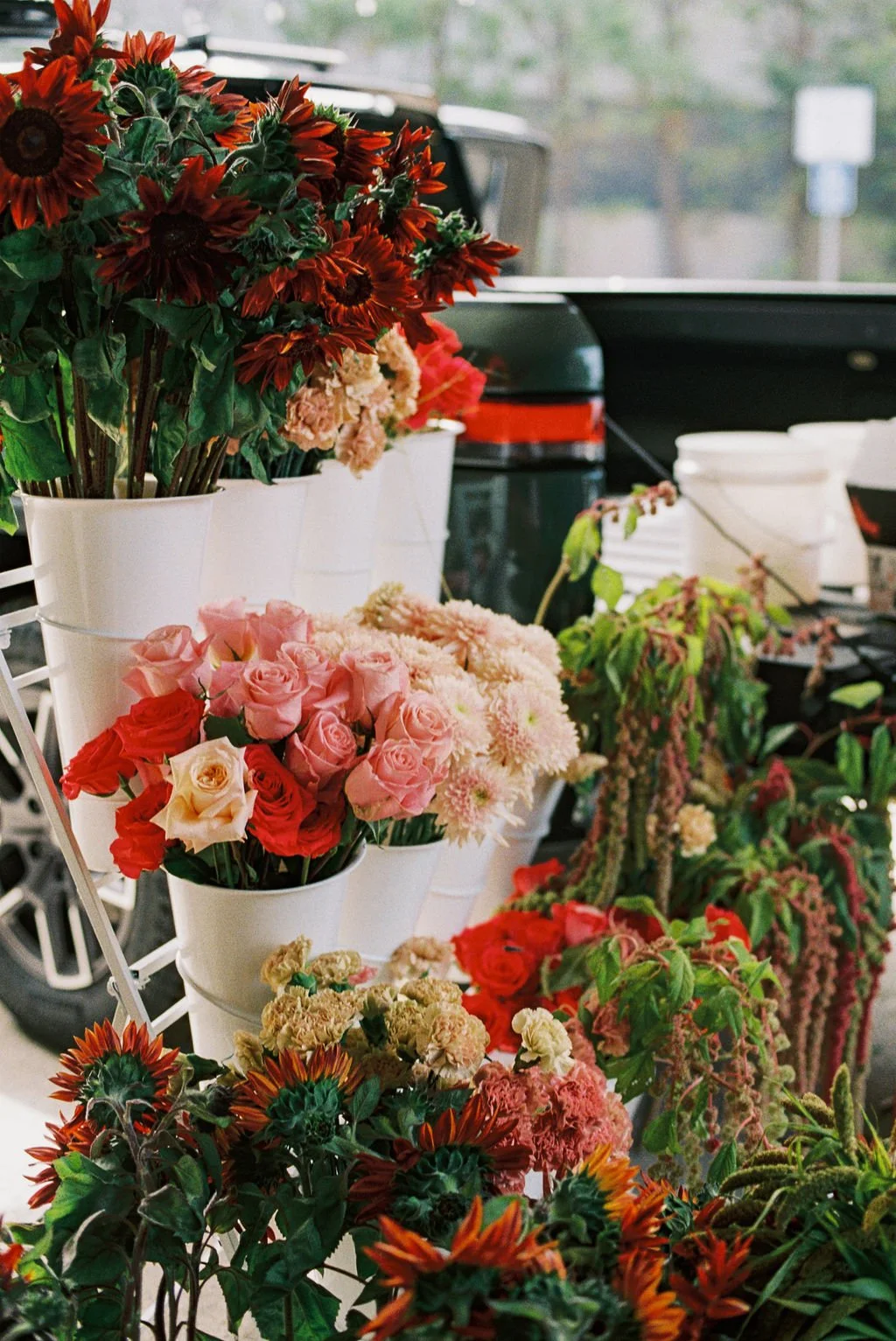 Various colorful flowers, including roses, sunflowers, and other blossoms, arranged in white buckets and on display, with a blurred background of a vehicle and trees.