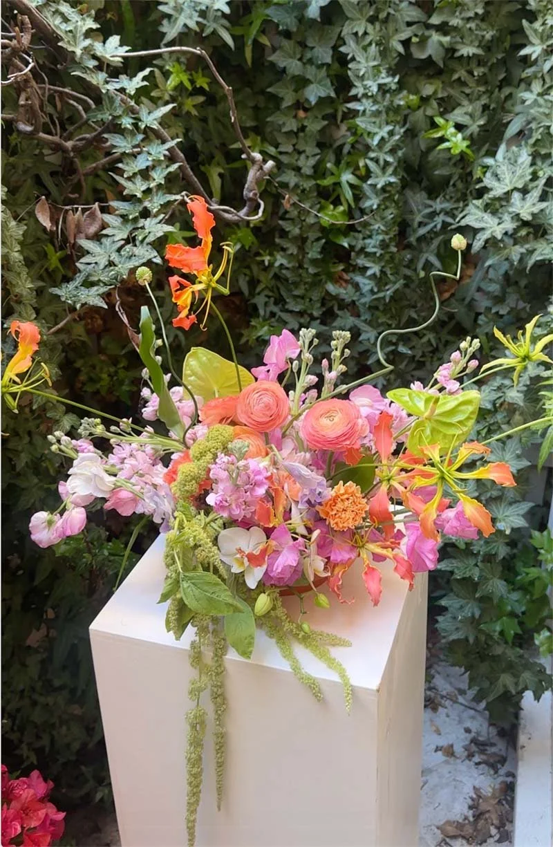 Colorful flower arrangement on a white pedestal, with pink, orange, and purple flowers, set against a green leafy background.