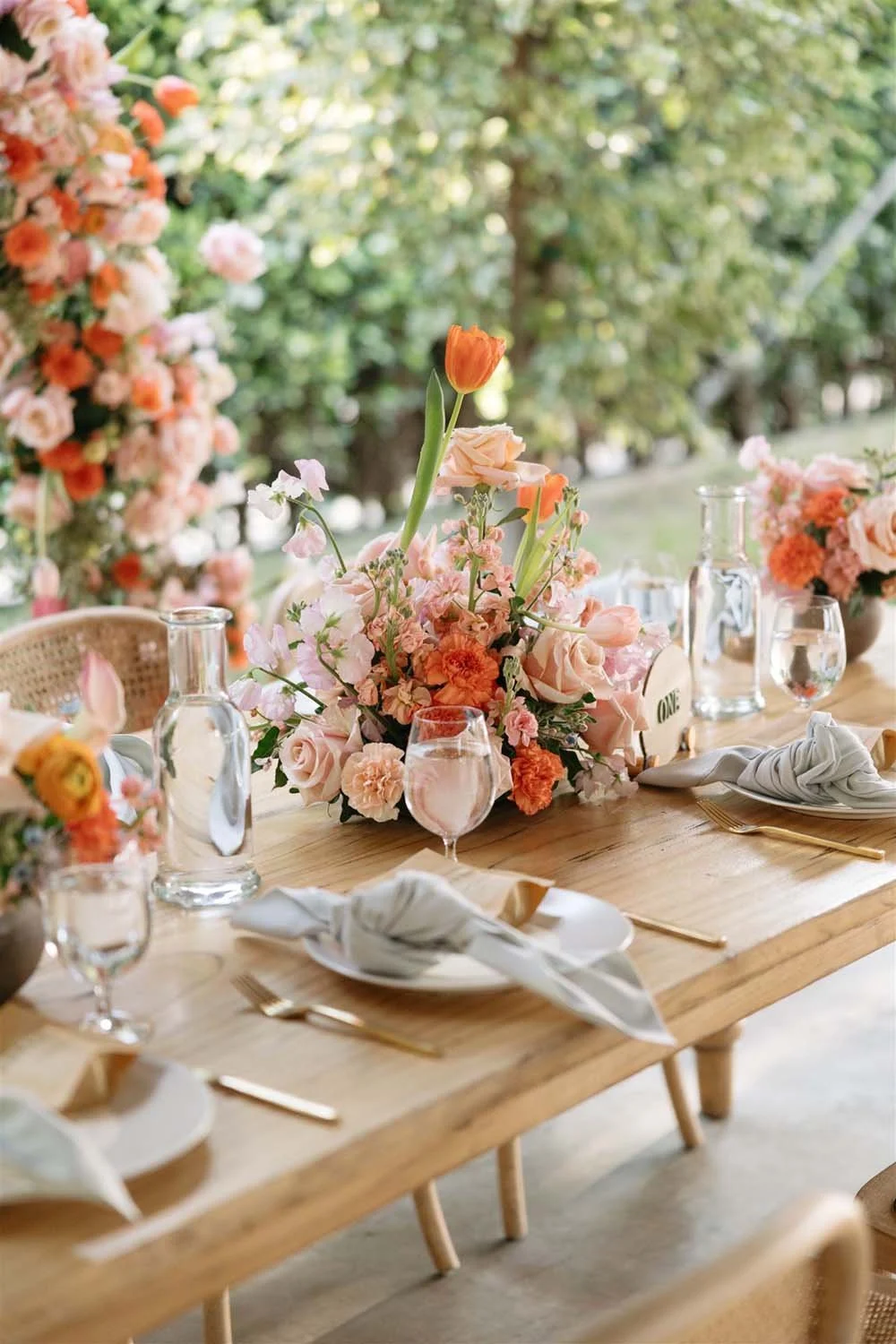 A decorated outdoor dining table with a floral centerpiece of pink, peach, and orange flowers, surrounded by water glasses, gold flatware, and white cloth napkins, with a lush green garden in the background.