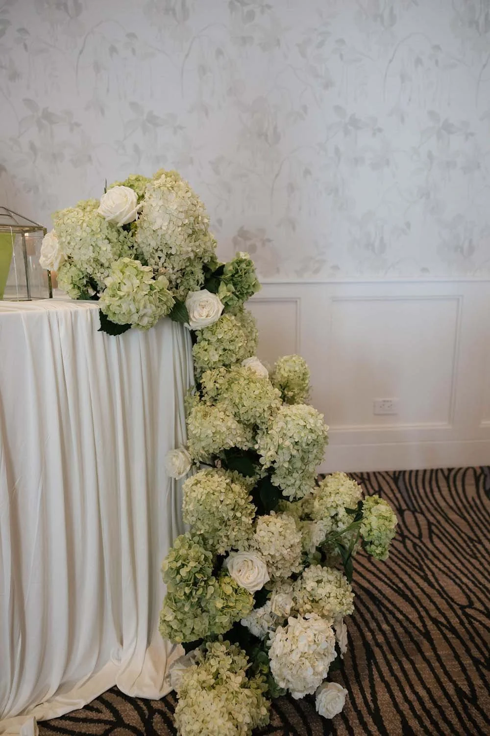 A cascading arrangement of white and cream hydrangeas and roses decorates a table with a white draped cloth, set against a neutral wall with floral wallpaper pattern.