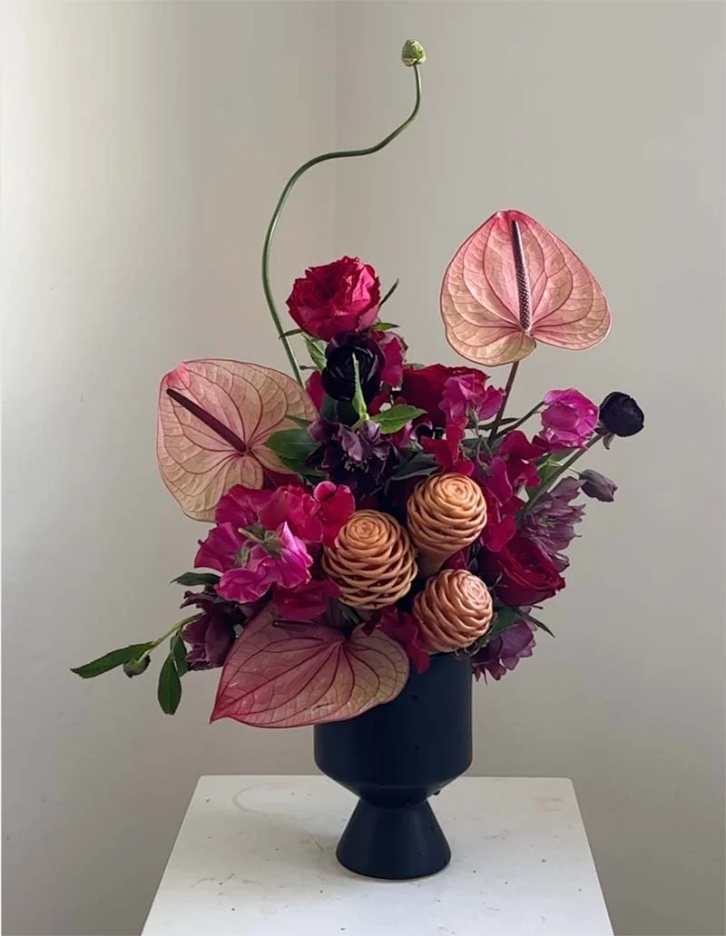 A floral arrangement in a black vase with pink, purple, and peach flowers, including pink anthuriums and pine cone-like flowers, accented with green leaves and a curved stem with a small flower at the top.