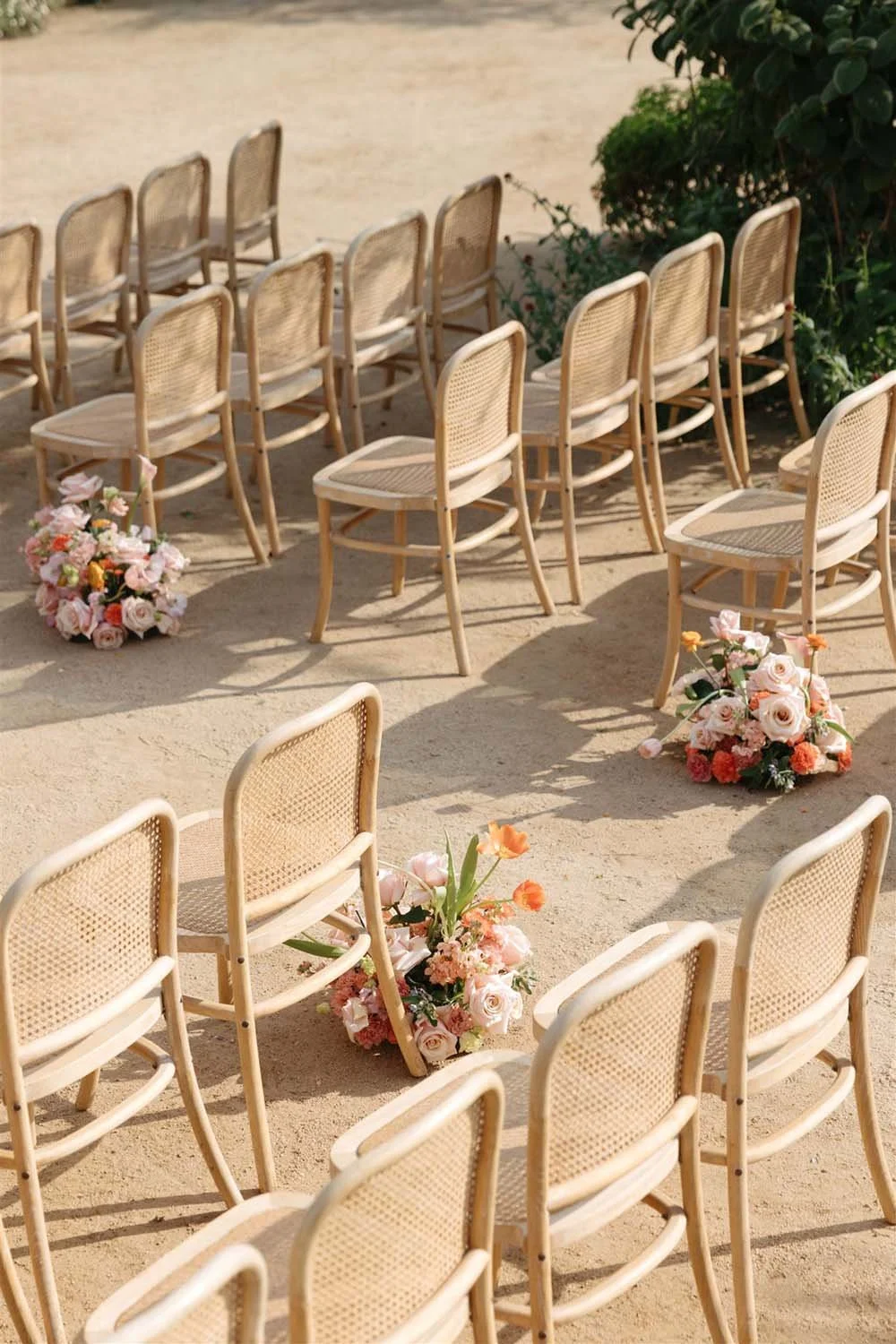 Outdoor wedding ceremony setup with multiple rows of beige chairs arranged on sand, decorated with bouquets of pink and peach flowers placed on the ground between the chairs.