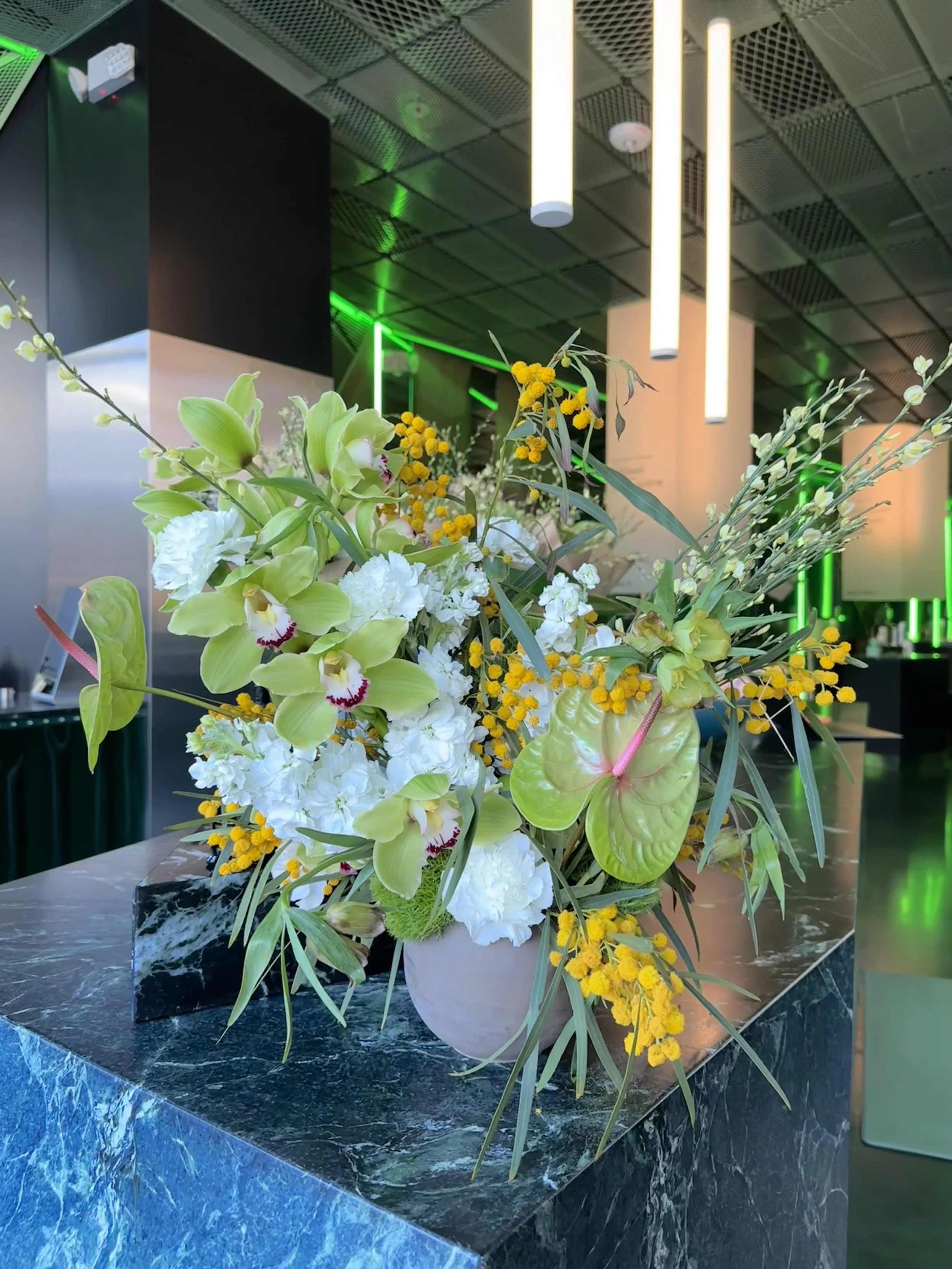 A colorful flower arrangement in a white vase on a black marble table in a modern interior with hanging lights and green accent lighting.