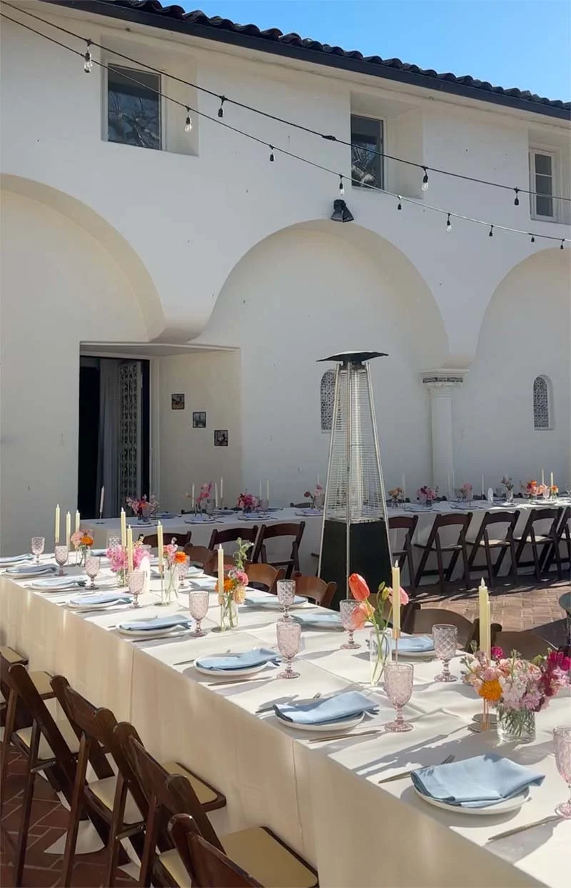 Long outdoor dining table set with plates, glasses, napkins, and floral centerpieces, under string lights, against a white building with arched doorways and small windows.
