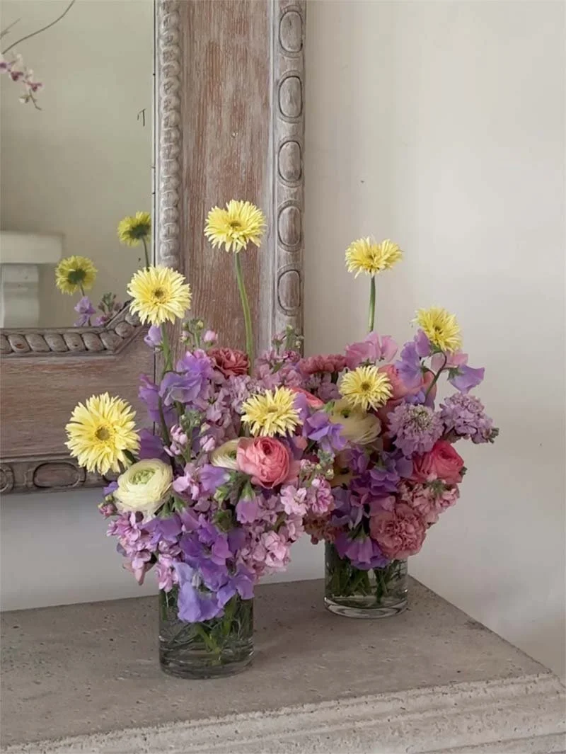 Two glass vases with colorful flowers including yellow, pink, purple, and white blooms, placed on a stone surface next to a rustic mirror frame and a mirror.