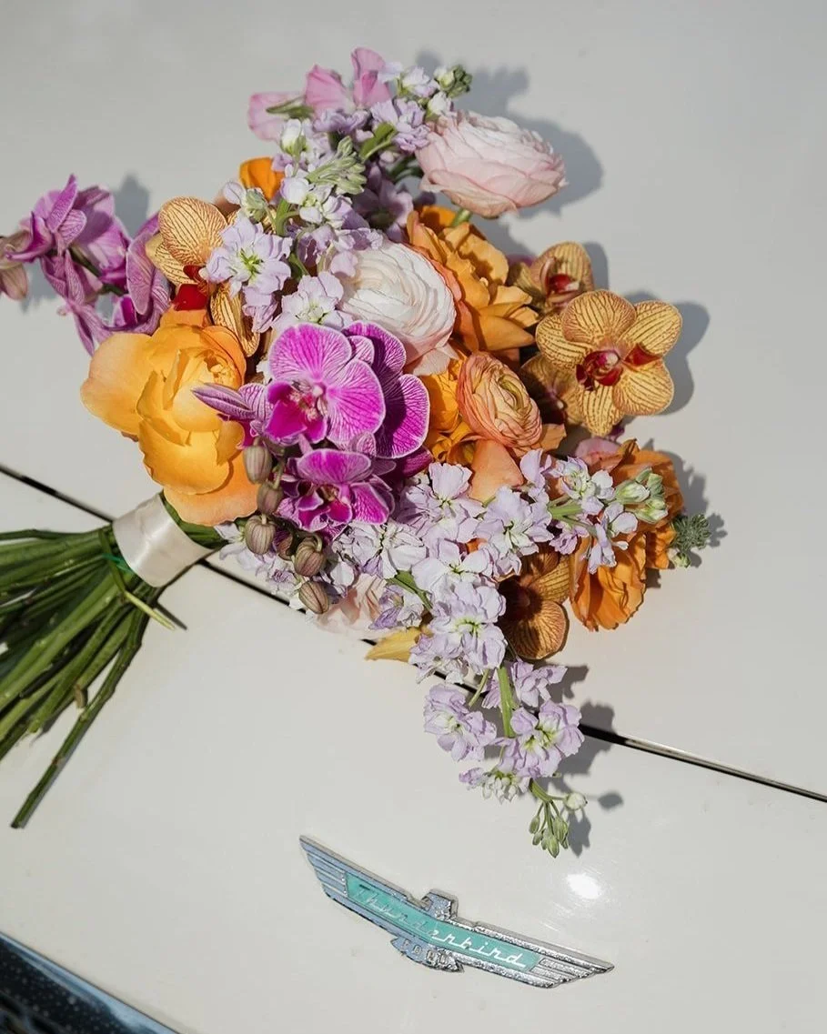 A colorful bouquet of mixed flowers, including orchids, ranunculus, and stock, placed on a white surface near a vintage Aston Martin badge.