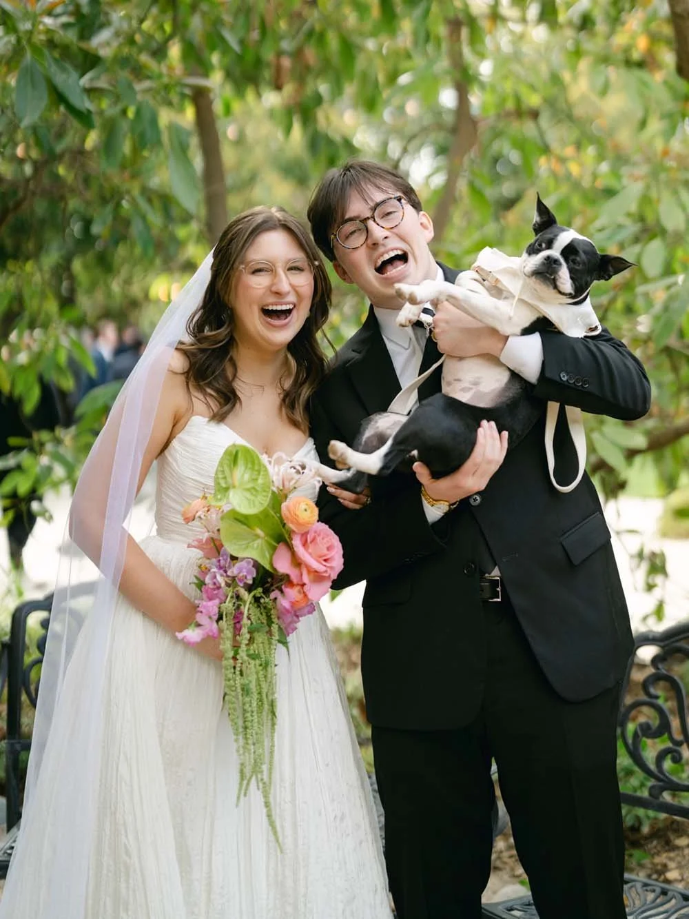 A joyful bride and groom smiling at the camera, holding a black and white dog, with the bride holding a bouquet of colorful flowers, outdoors with green trees in the background.