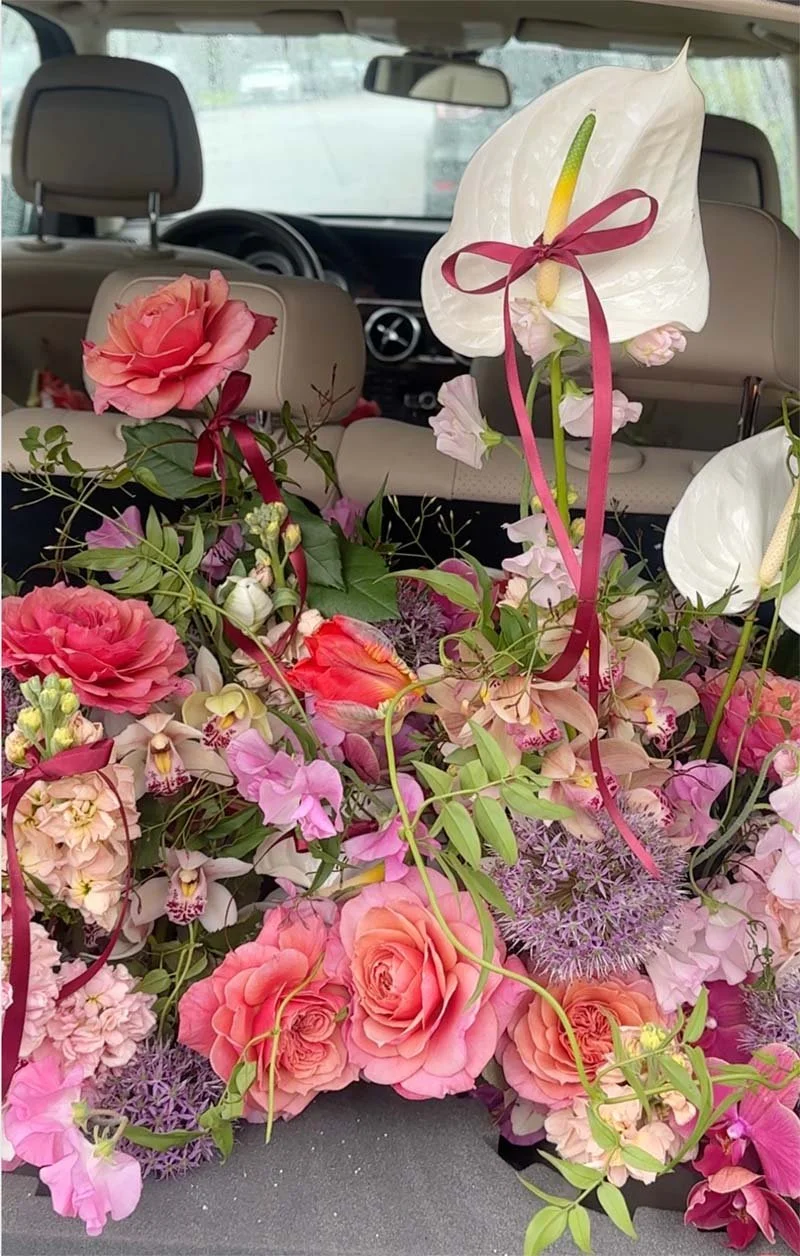 A large bouquet of pink, white, and peach flowers, including roses, anthuriums, and other blooms, inside a vehicle. The flowers are decorated with pink ribbons and arranged on a car seat.