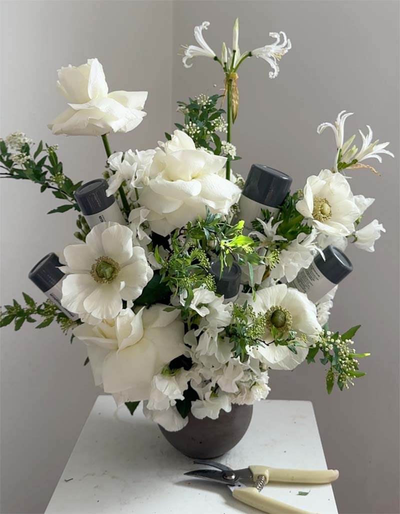 A bouquet of white flowers, including roses, anemones, and lilies, in a dark vase with gardening pruning shears resting nearby on a white surface.