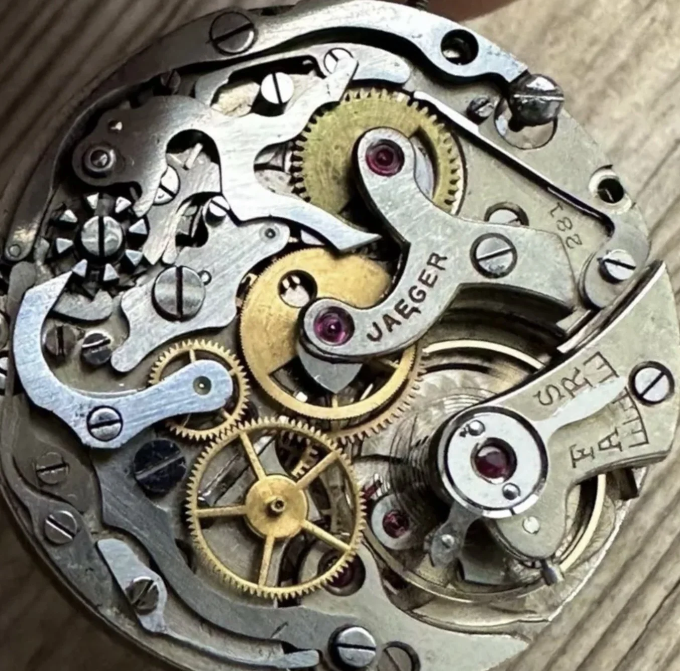 Close-up of the inner mechanical gears and components of a watch movement, showing metal gears, screws, and a bridge engraved with the brand name 'JAEGER'.