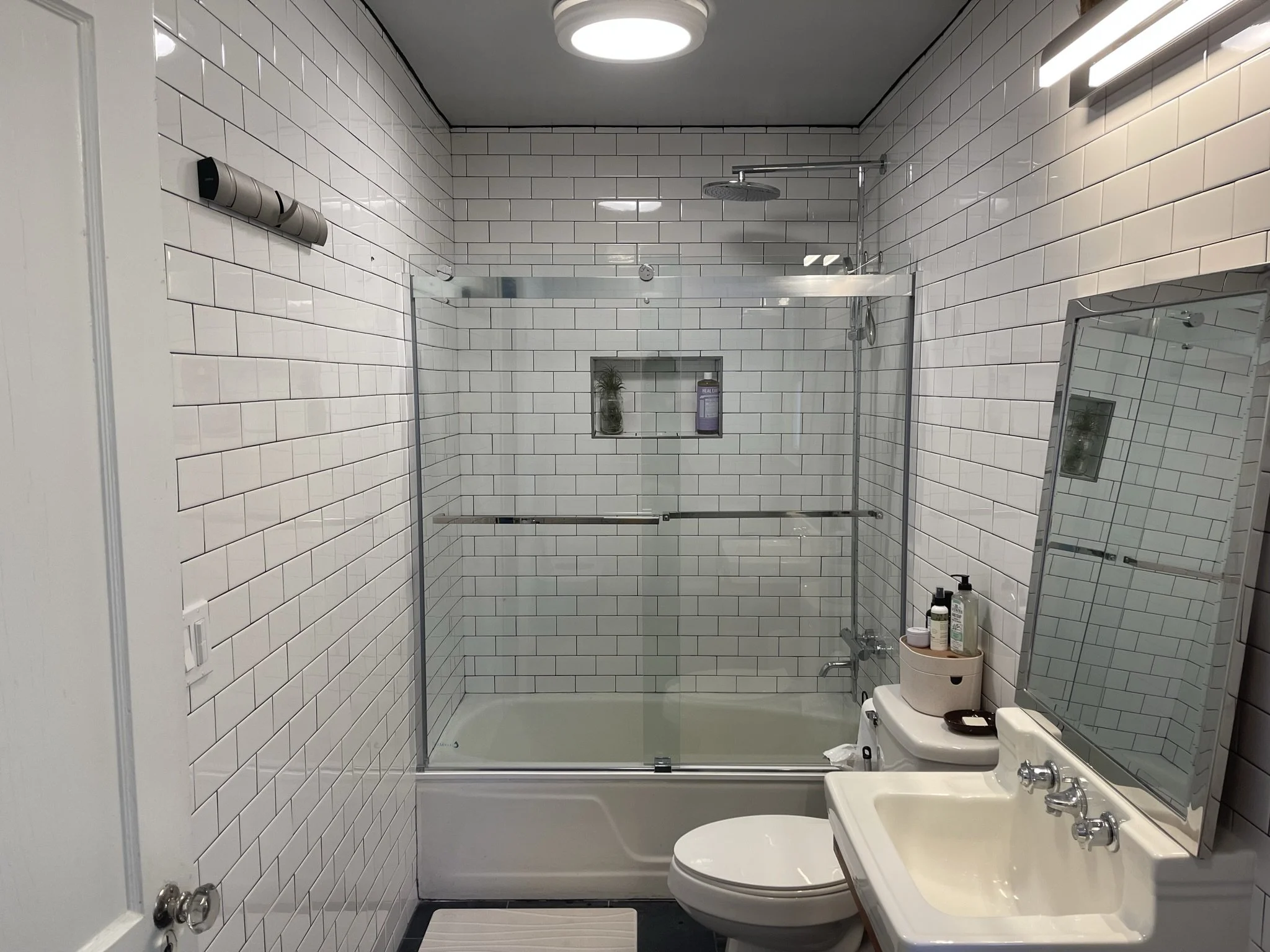 Modern bathroom renovation in West Asheville featuring white subway tile walls with black grout, dark slate floor tiles, and a sliding glass bathtub door. The design includes a vintage-style wall-mount sink and a recessed shower niche, blending indus