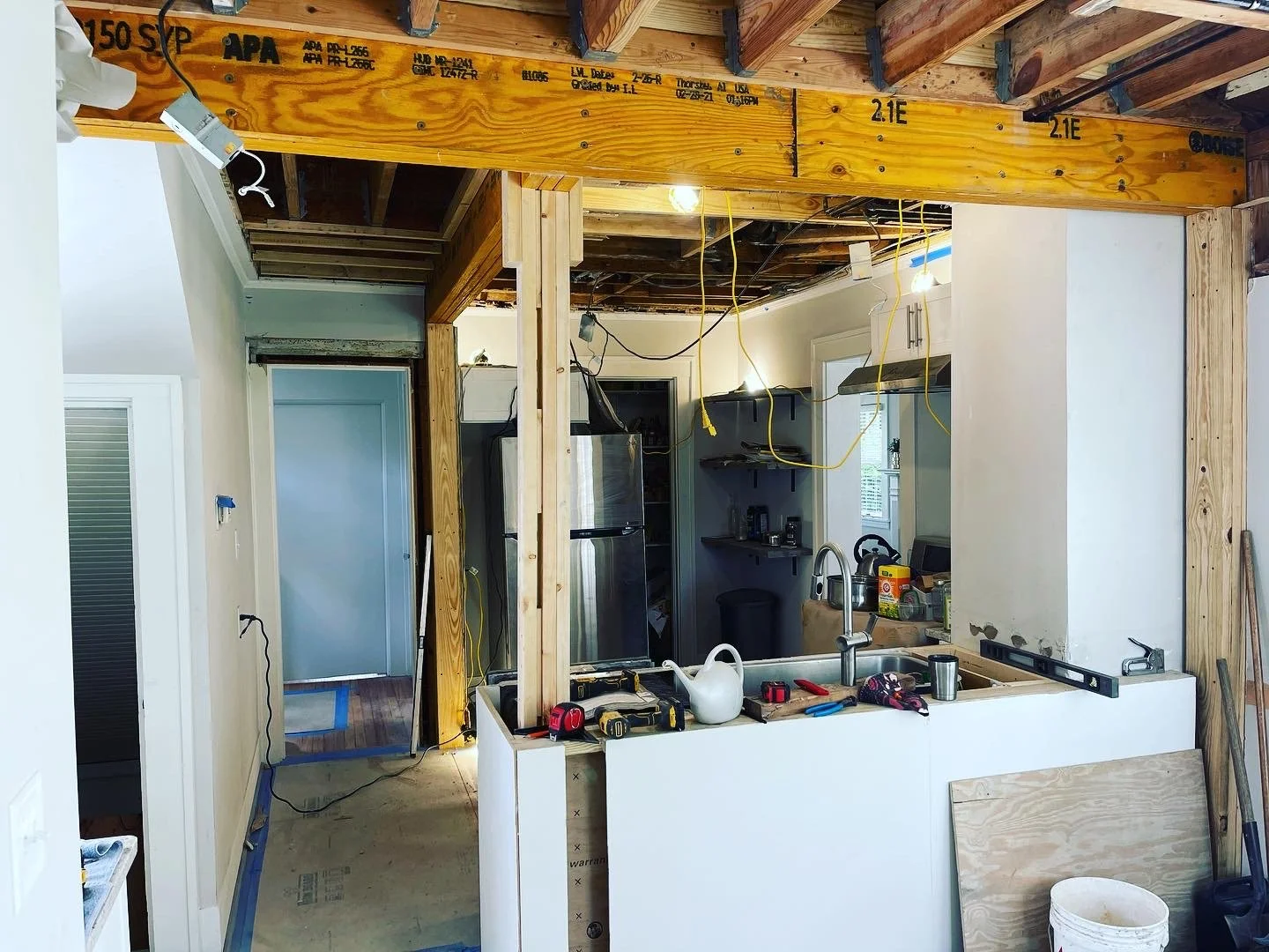 A detailed 'before' snapshot of a structural renovation in West Asheville, where a load-bearing wall has been removed to open up a kitchen. The image prominently shows a large laminated veneer lumber (LVL) support beam and exposed posts essential for
