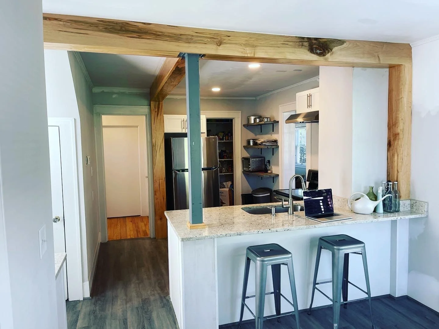 The 'after' result of an open concept kitchen renovation in West Asheville, featuring a breakfast bar with granite countertops and industrial metal stools. The structural support is now finished with beautiful natural wood-wrapped beams and a teal ac
