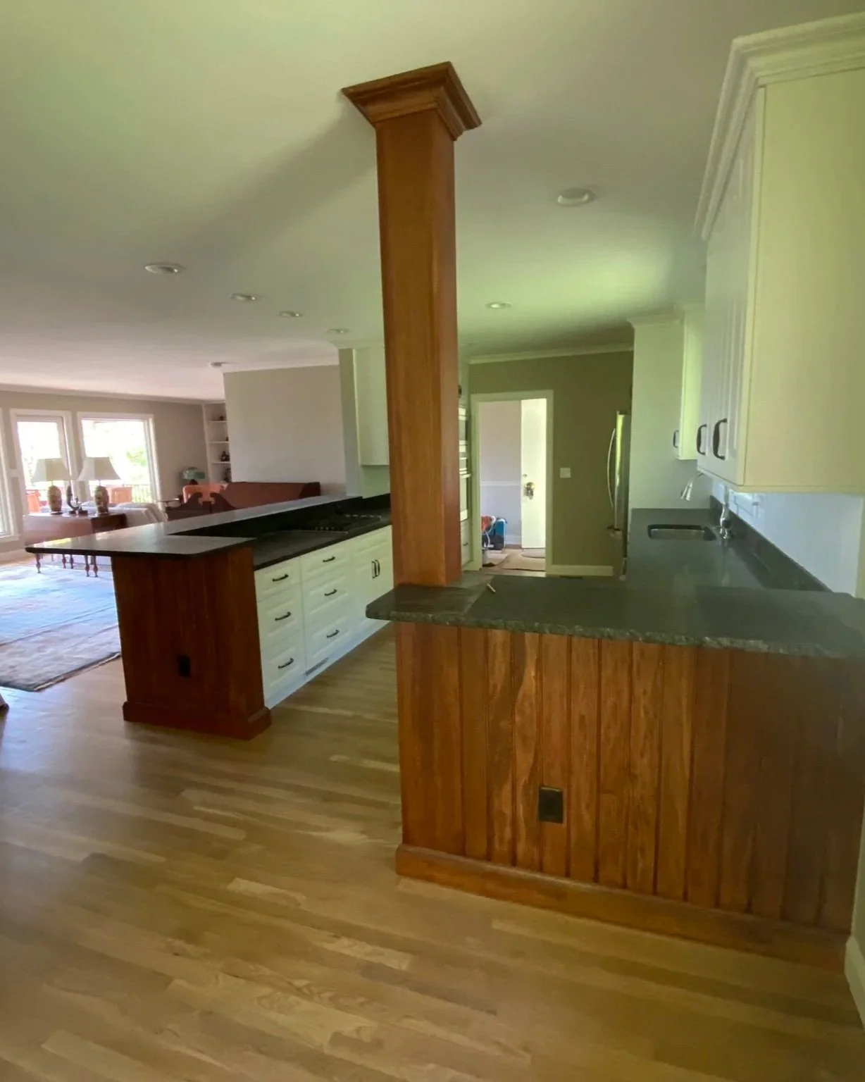 Open-concept kitchen remodel in Asheville, NC, featuring a custom wood-paneled peninsula with black soapstone countertops, white cabinetry, and a matching wood structural support column on refinished oak floors.