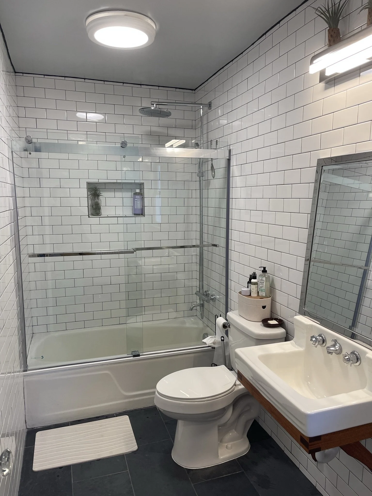 Modern bathroom renovation in West Asheville featuring white subway tile walls with black grout, dark slate floor tiles, and a sliding glass bathtub door. The design includes a vintage-style wall-mount sink and a recessed shower niche, blending indus