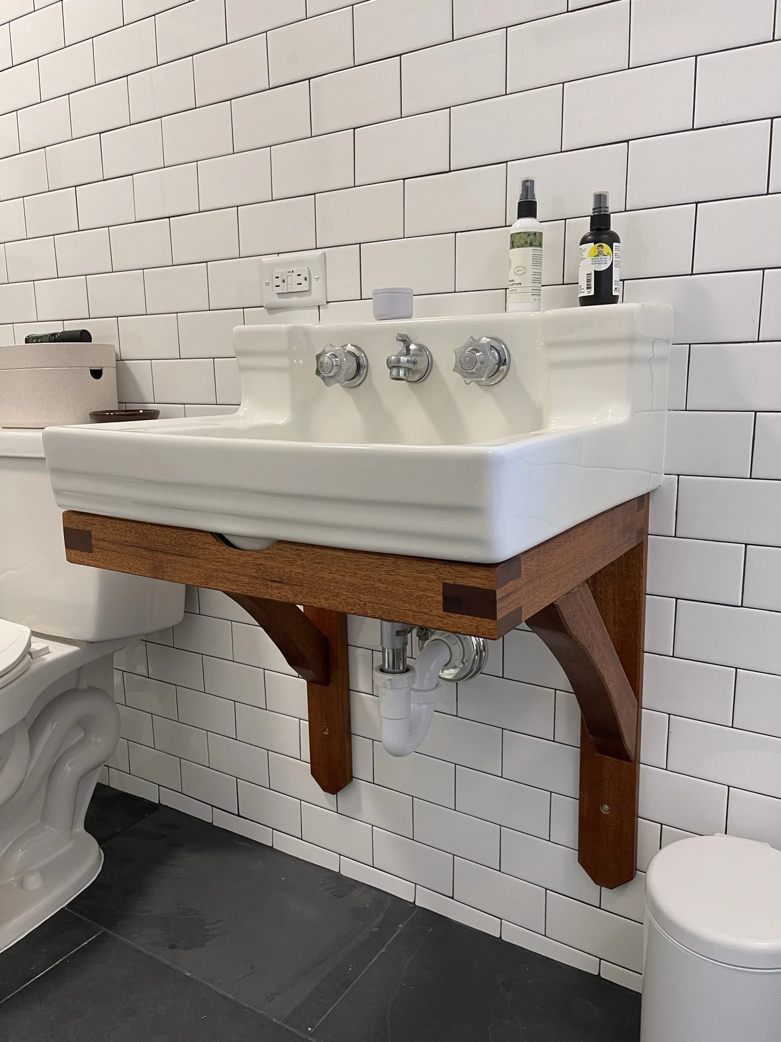 Close-up of a custom wall-mounted vintage sink with a handcrafted wood support frame in a West Asheville bathroom. The design features white subway tile with dark grout and slate floor tiles, showcasing a blend of mid-century and modern industrial st