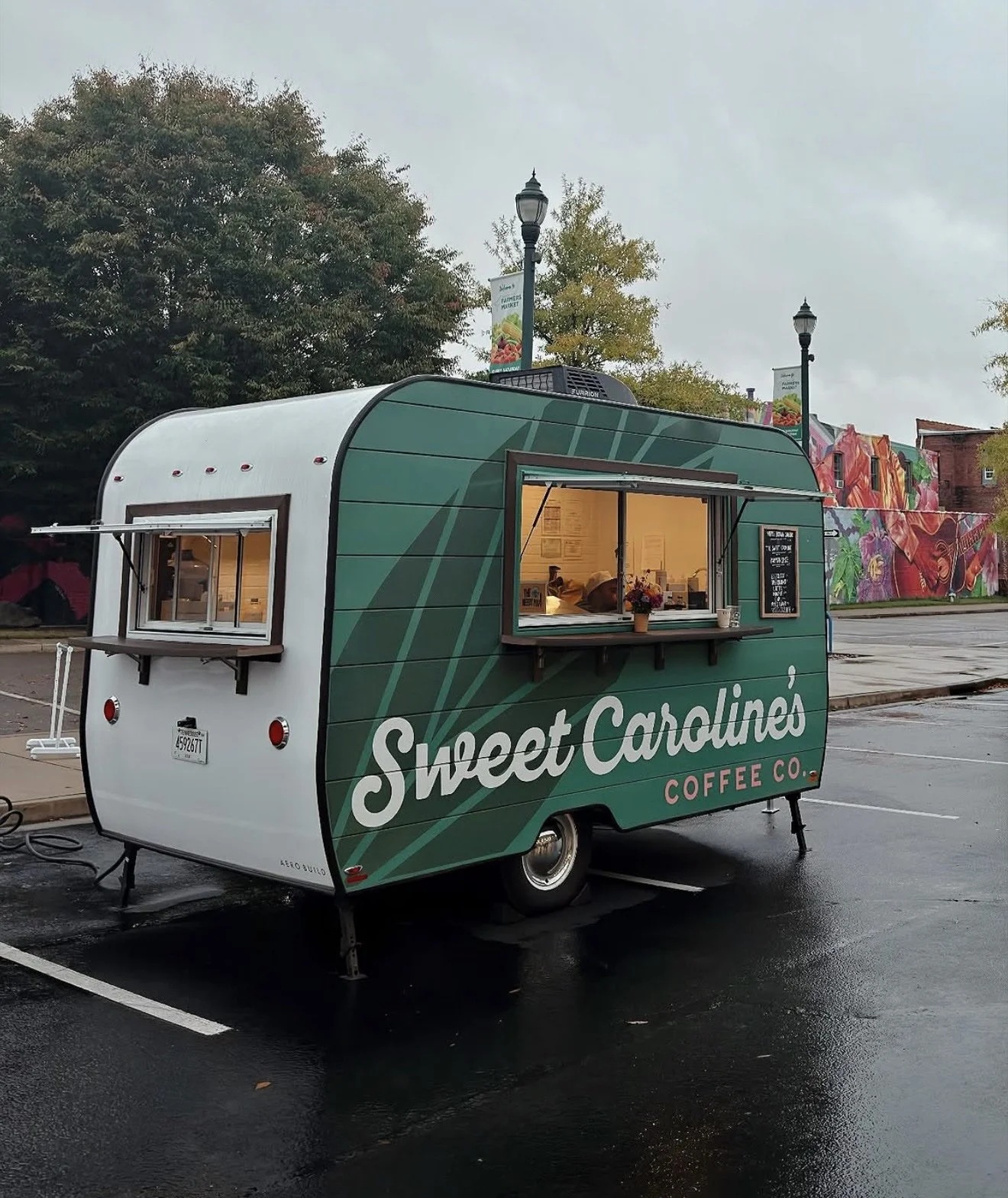 A small green food truck named Sweet Caroline's Coffee Co. is parked in a parking lot with street lamps and trees in the background. The truck has a serving window with a person inside and a small chalkboard menu outside.
