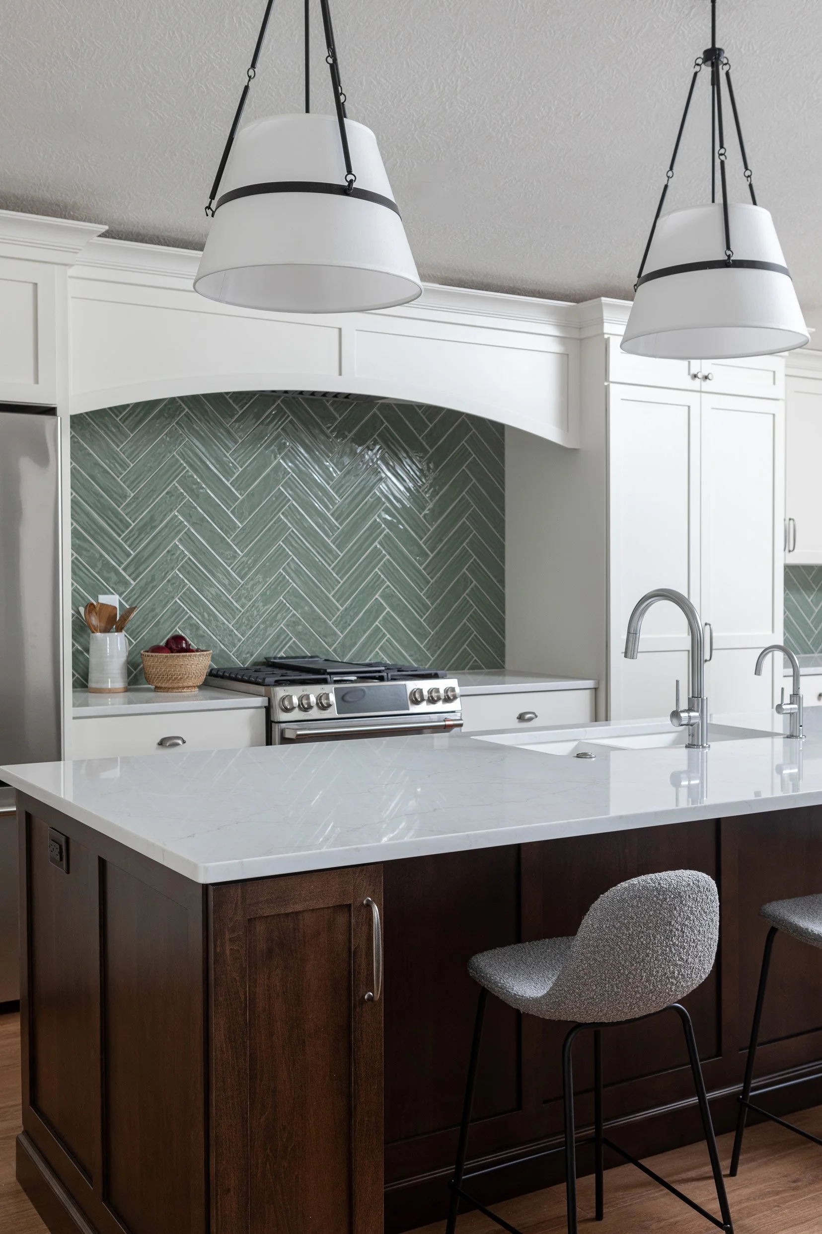 Modern kitchen with a white marble island, dark wood base, two gray barstools, green herringbone tile backsplash, white cabinets, stainless steel stove, and two white pendant lights hanging from the ceiling.