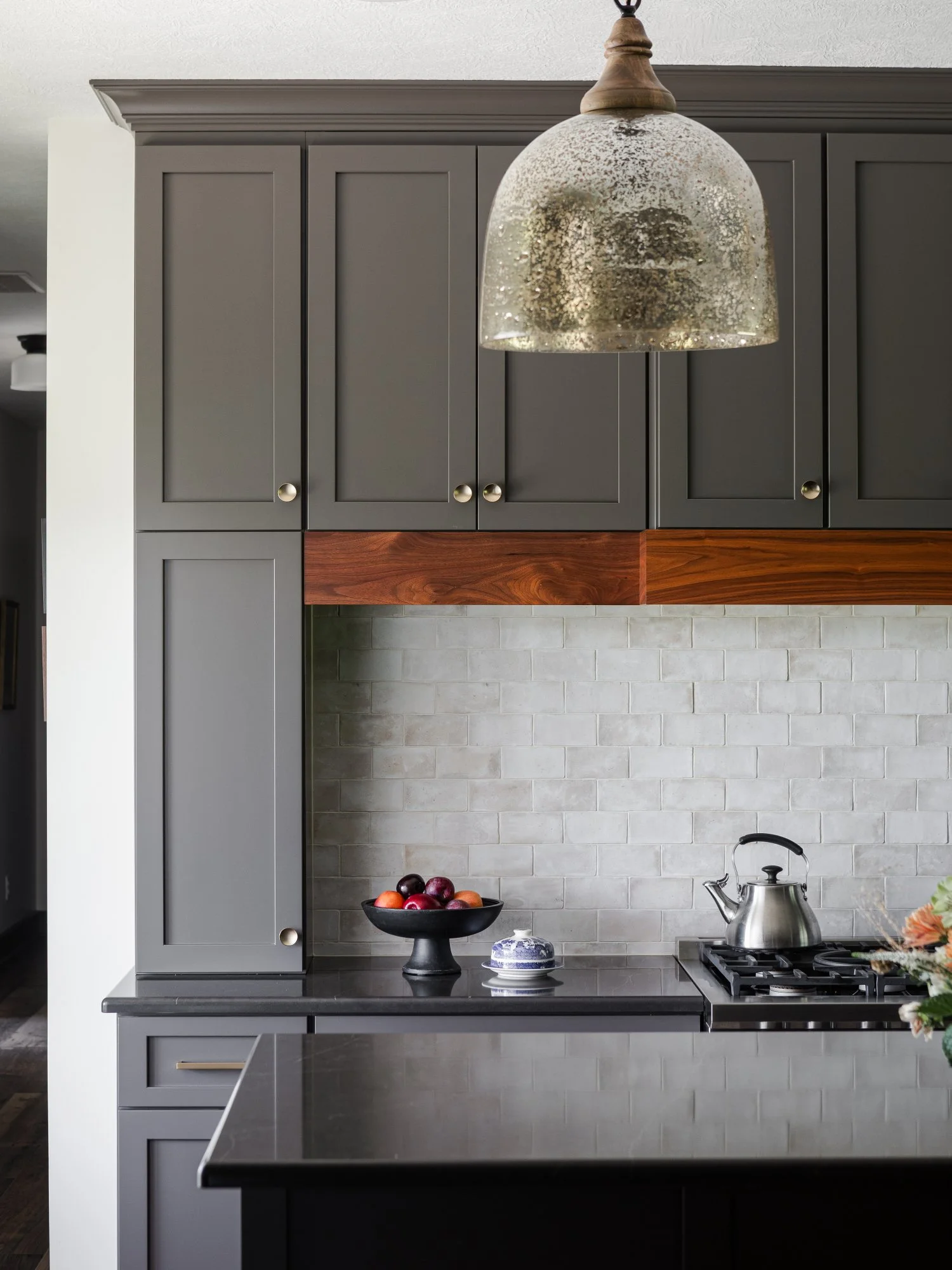 Kitchen with gray cabinets, a black countertop, a silver teapot on the stove, a black bowl with apples, and a glass covered dish on the counter. A textured glass pendant light hangs above.