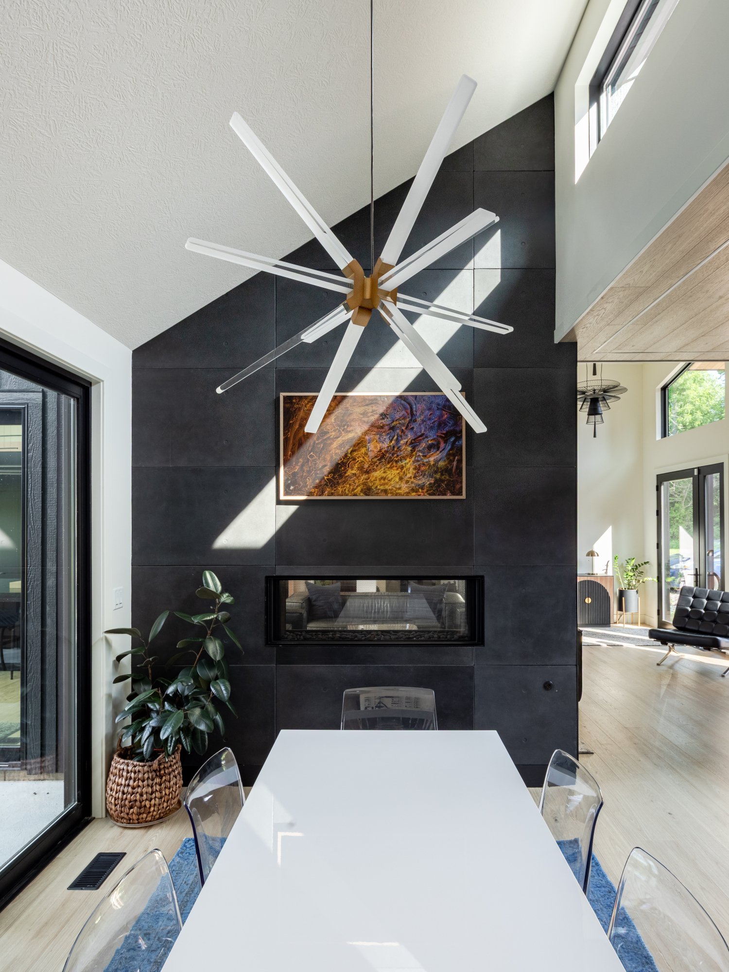 Modern dining area with white table, transparent chairs, black tiled wall with fireplace, abstract artwork, large window, and high ceiling with unique chandelier