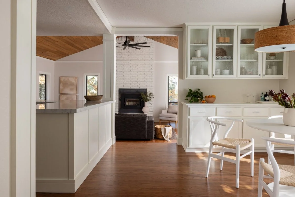 Open-concept kitchen with white cabinets, a dining table with white chairs, and a living area with a white brick fireplace and a dark sofa.
