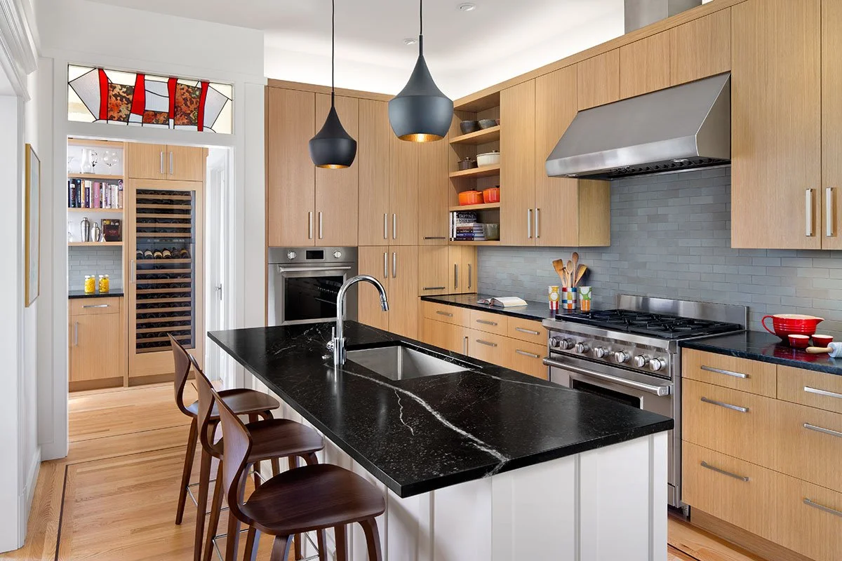 Modern kitchen with black island countertop, light wood cabinets, stainless steel appliances, hanging pendant lights, and a wine fridge in the background.