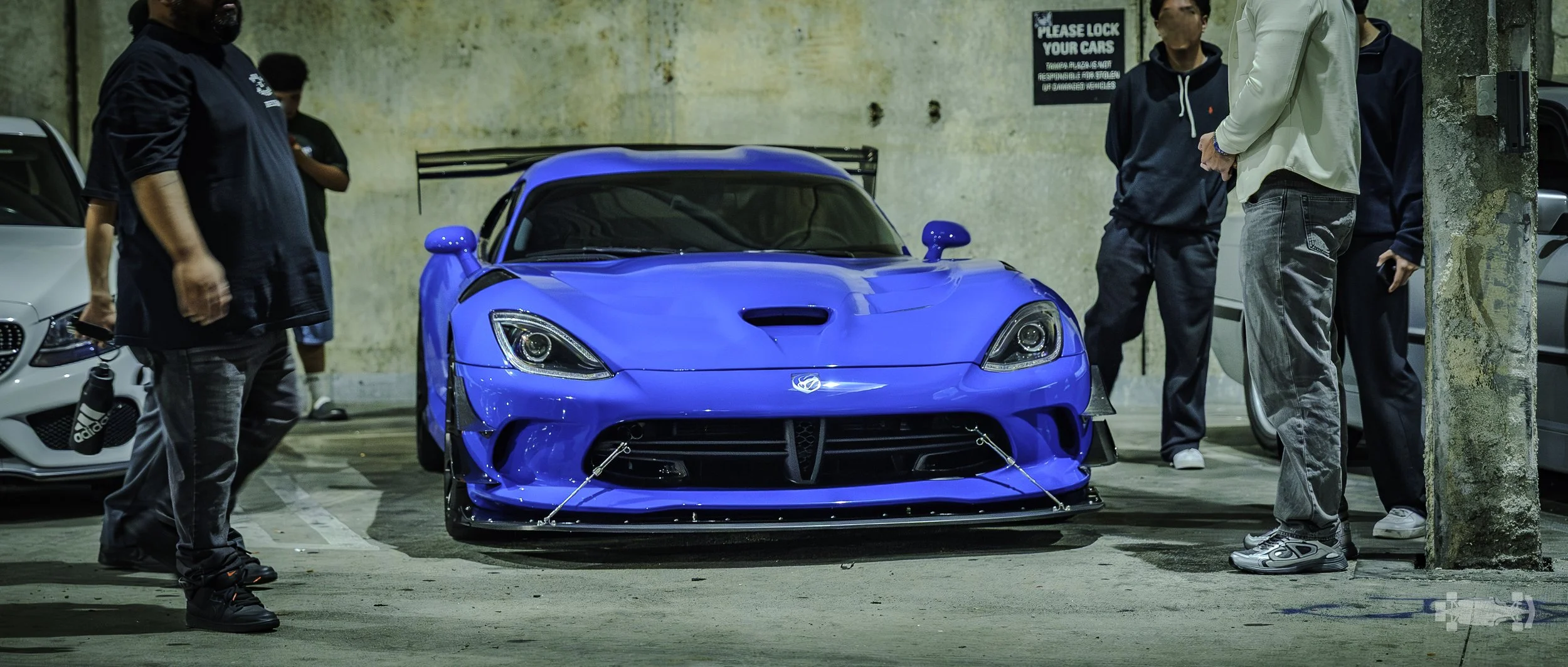 A blue racing car with a spoiler and front splitters is parked in an indoor parking garage, surrounded by several people.