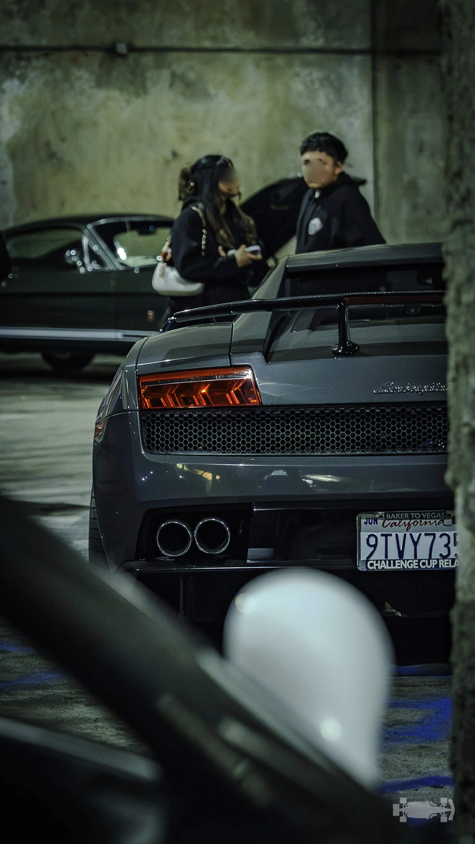 A gray sports car with a spoiler and dual exhaust pipes parked in an underground parking garage, with two people standing nearby, one looking at a phone. The car has a California license plate.