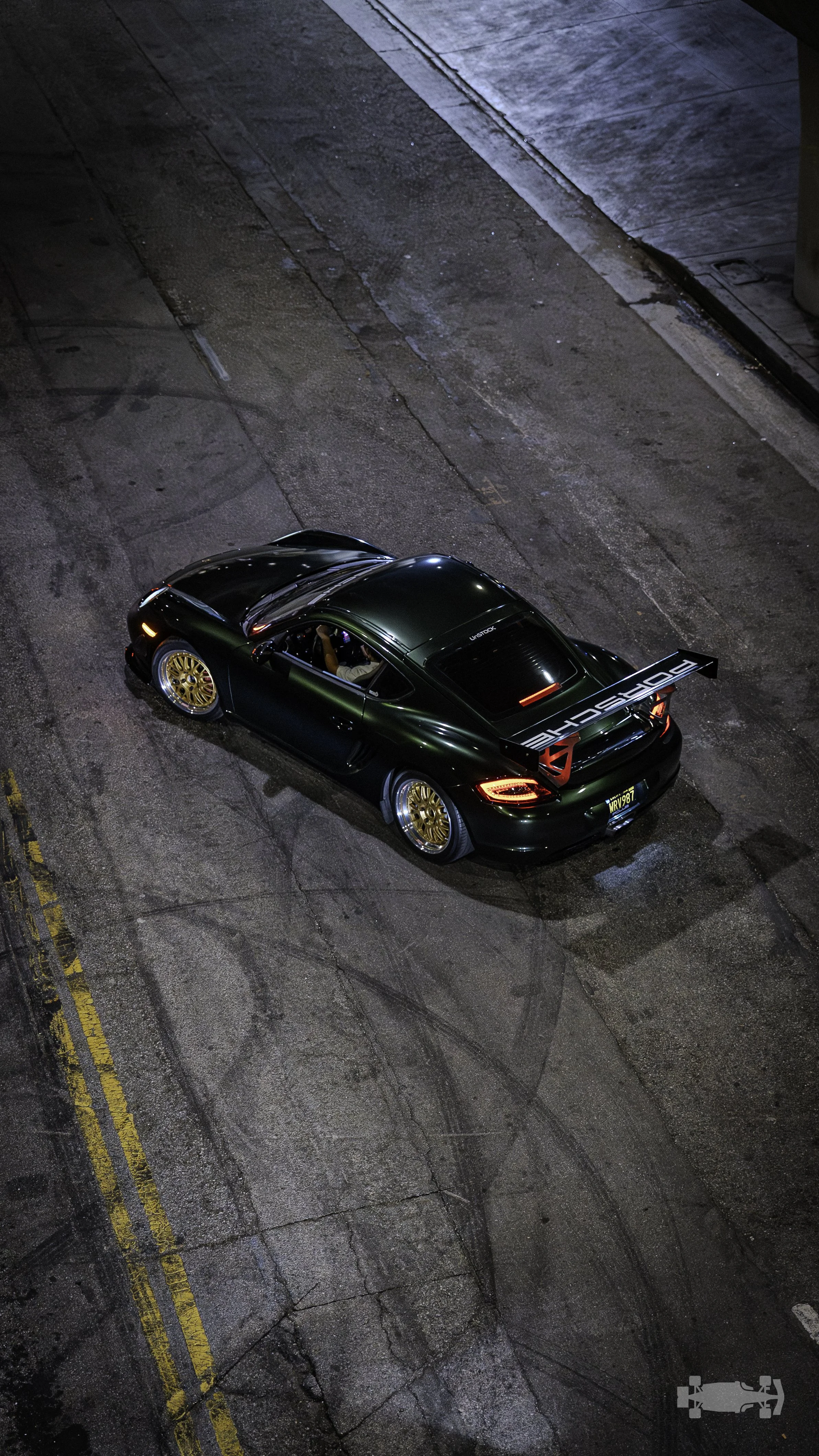 A dark green sports car with a large rear wing, gold wheels, and racing decals, parked on a street at night.